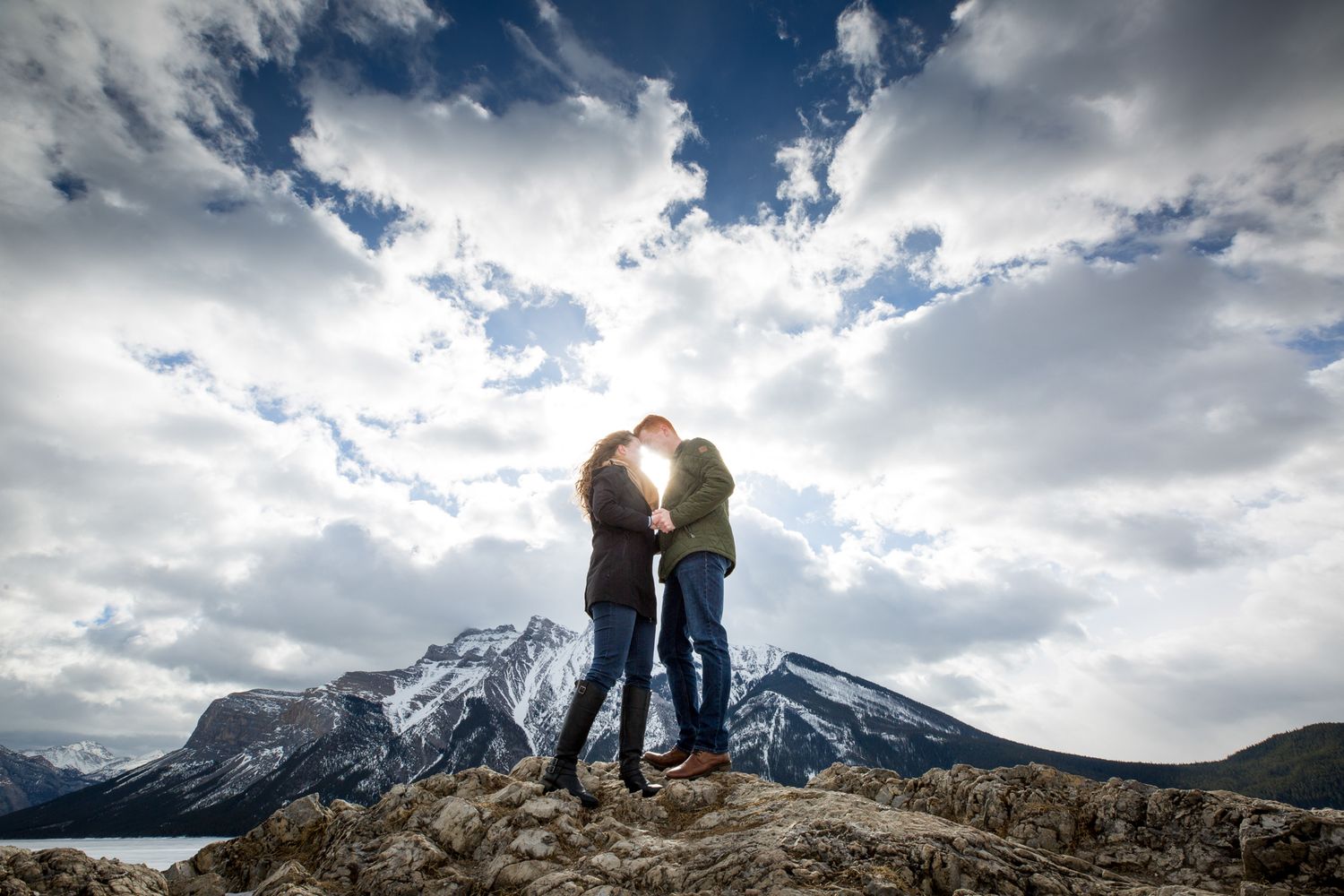 Couple kissing on mountain summit with dramatic cloudy sky and snow-capped peaks in background at Lake Minnewanka in Banff National Park.
