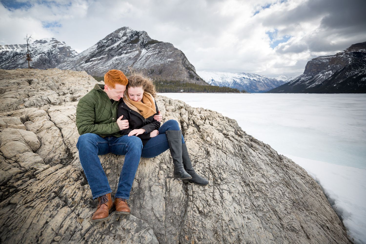 Couple sitting together on rocky shore with frozen lake and snow-capped mountains in background at Lake Minnewanka in Banff National Park.