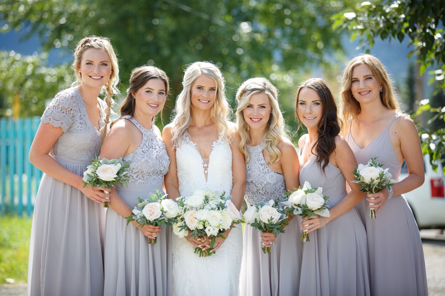 Bridesmaids wearing light gray dresses pose together holding white floral bouquets in an outdoor setting. Quarry lake park in Canmore.