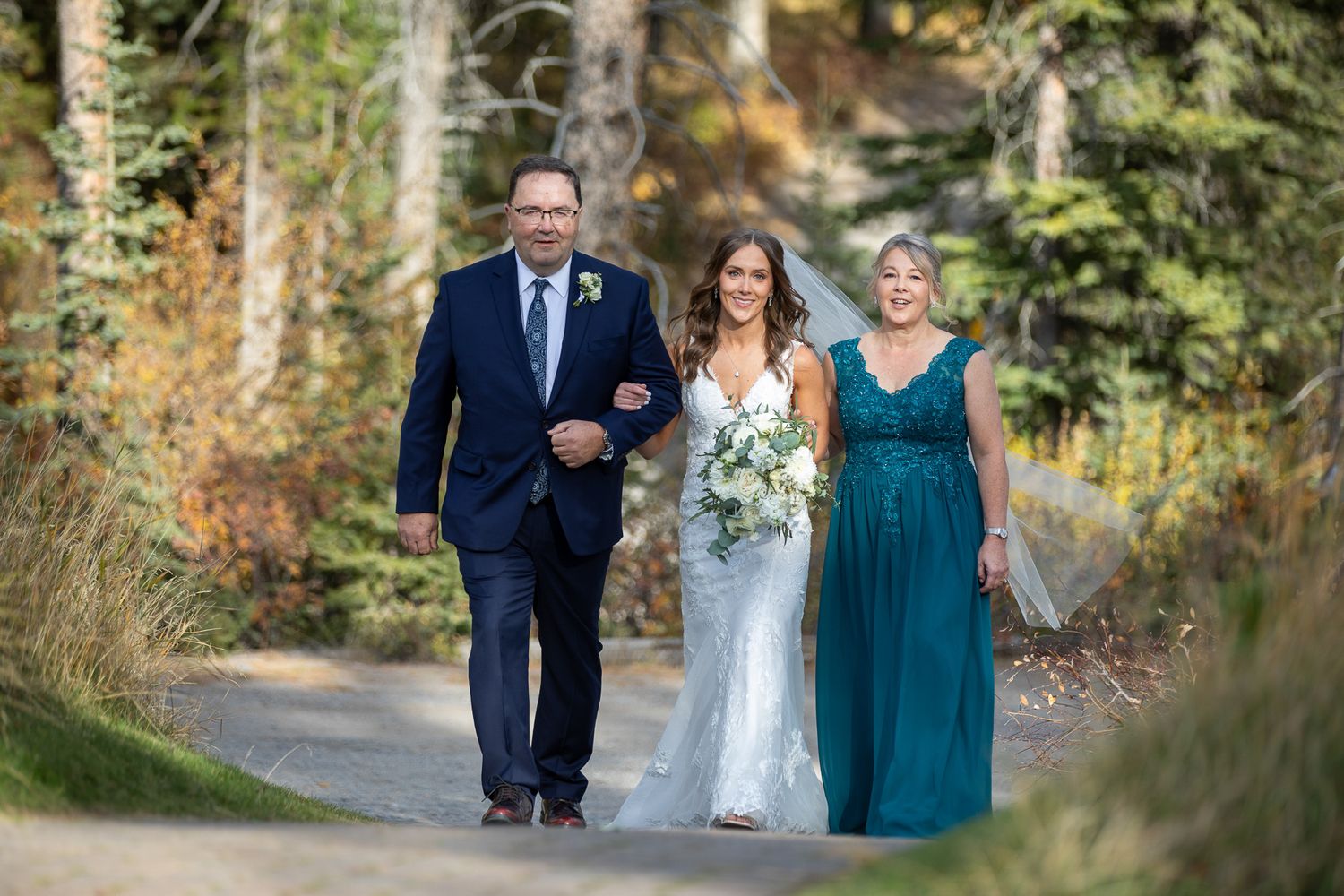 Wedding party walking along a wooded path in autumn with bridesmaids wearing teal dresses at Silvertip resort gazebo in Canmore, AB.