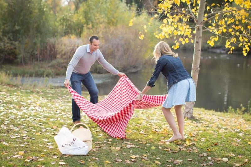 Couple spreading a red checkered blanket on grass for an autumn picnic by a lake at Fish Creek Park in Calgary.