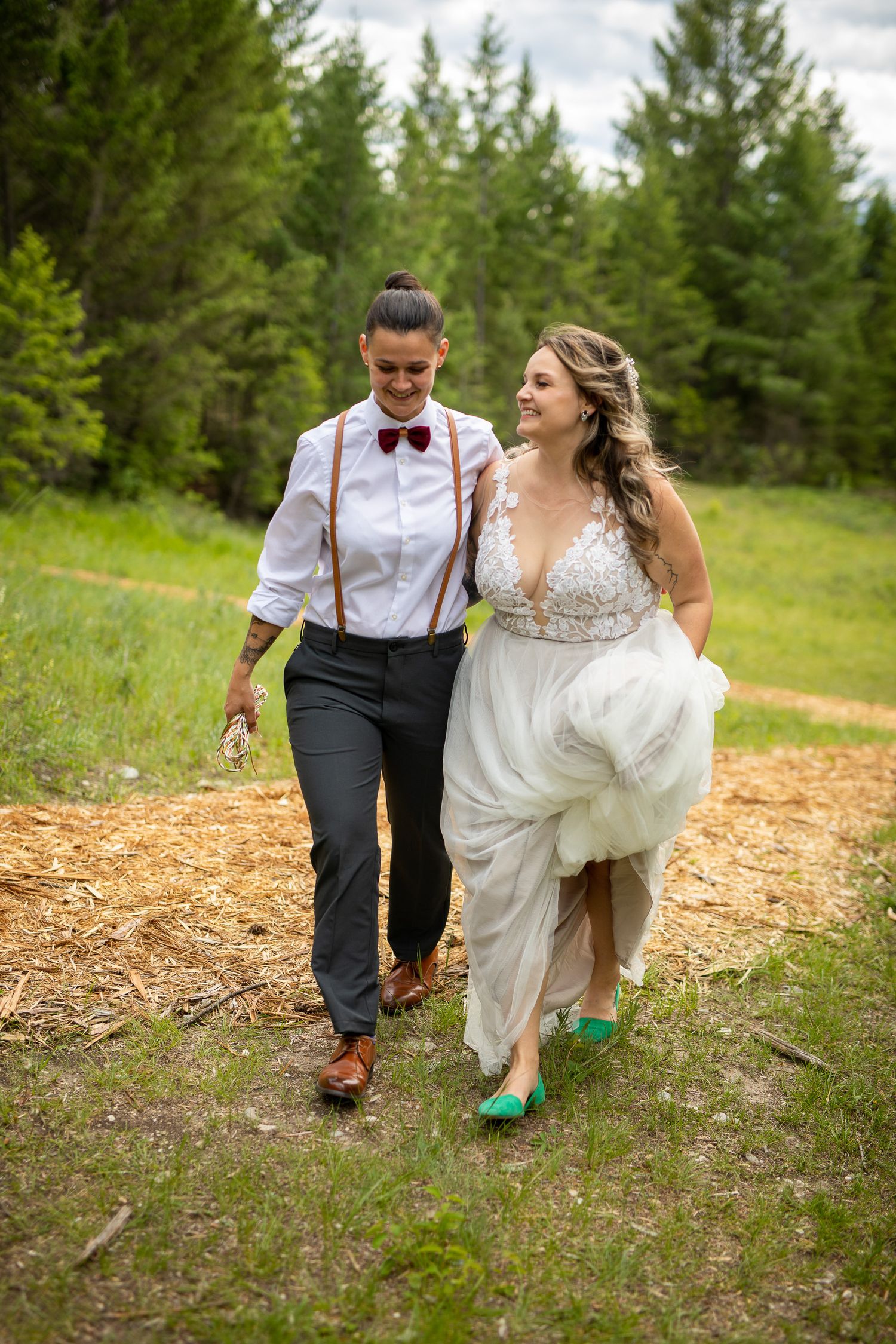 Two brides walks together on a dirt path through evergreens, wearing wedding attire with suspenders and green shoes. Radium, BC.