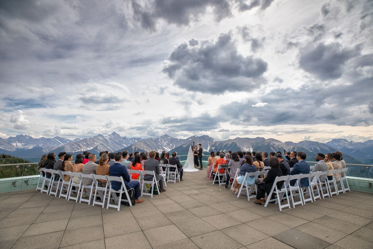 A panoramic sequence of a scenic mountain wedding ceremony with guests seated under dramatic cloudy skies at Sky Bistro terrace, Banff AB.