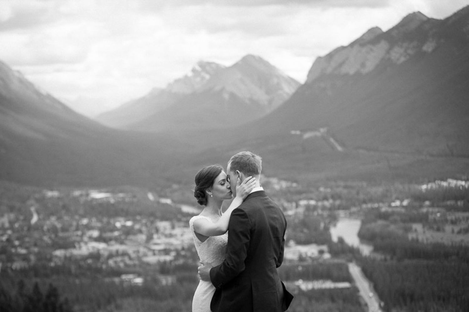 A couple embraces while overlooking a scenic mountain landscape with dramatic peaks and valleys in black and white. Norquay Lookout in Banff