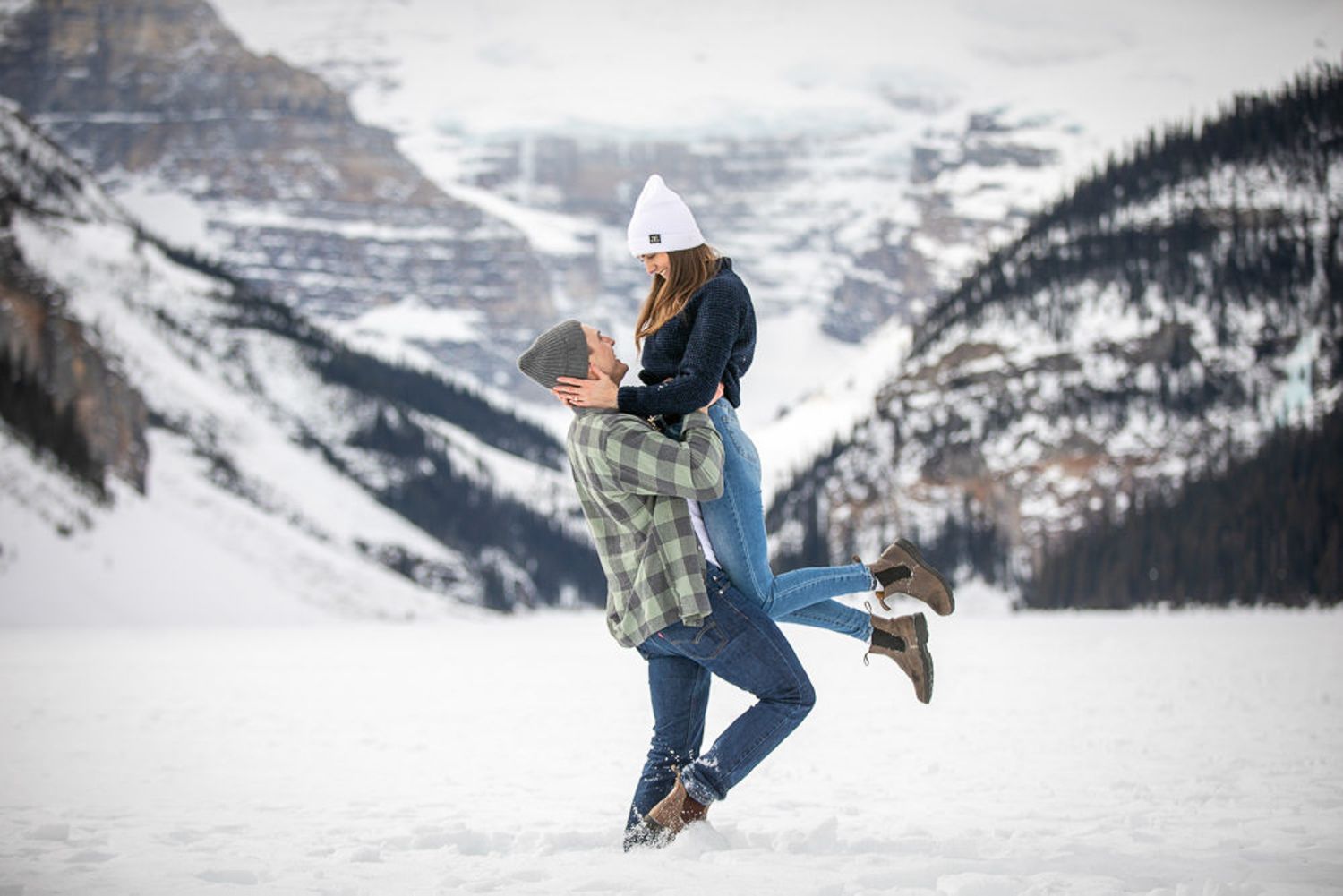 A couple embraces in winter attire against a snowy mountain backdrop at Lake Louise in the Canadian Rockies.