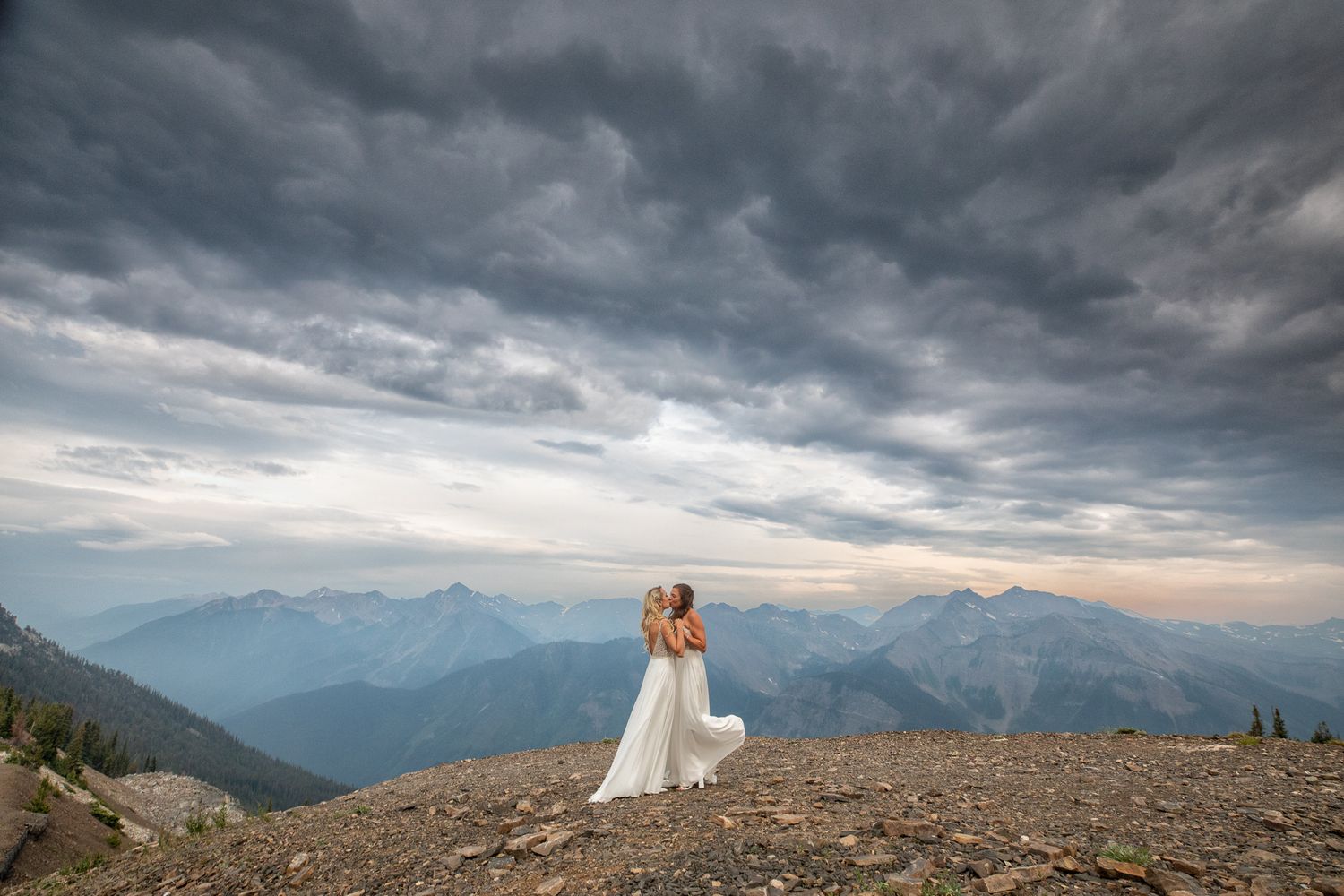 Bride in white gown stands on rocky mountain summit embracing partner under dramatic stormy sky with alpine peaks behind at Kicking Horse Re