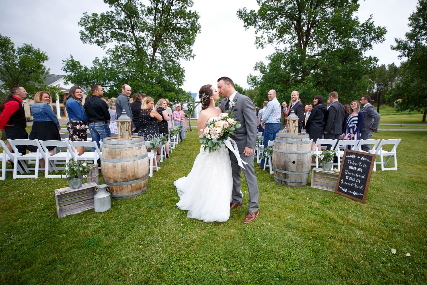 Bride and groom share a kiss at outdoor wedding ceremony at Cherry creek estate on green lawn with barrel decorations and seated guests