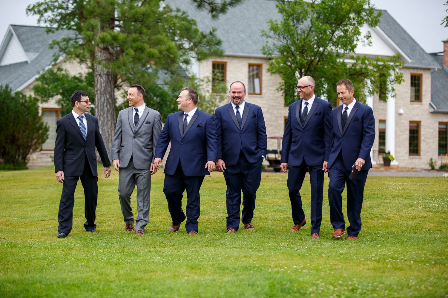 Six groomsmen in navy and gray suits walking together on a green lawn in front of an elegant estate.