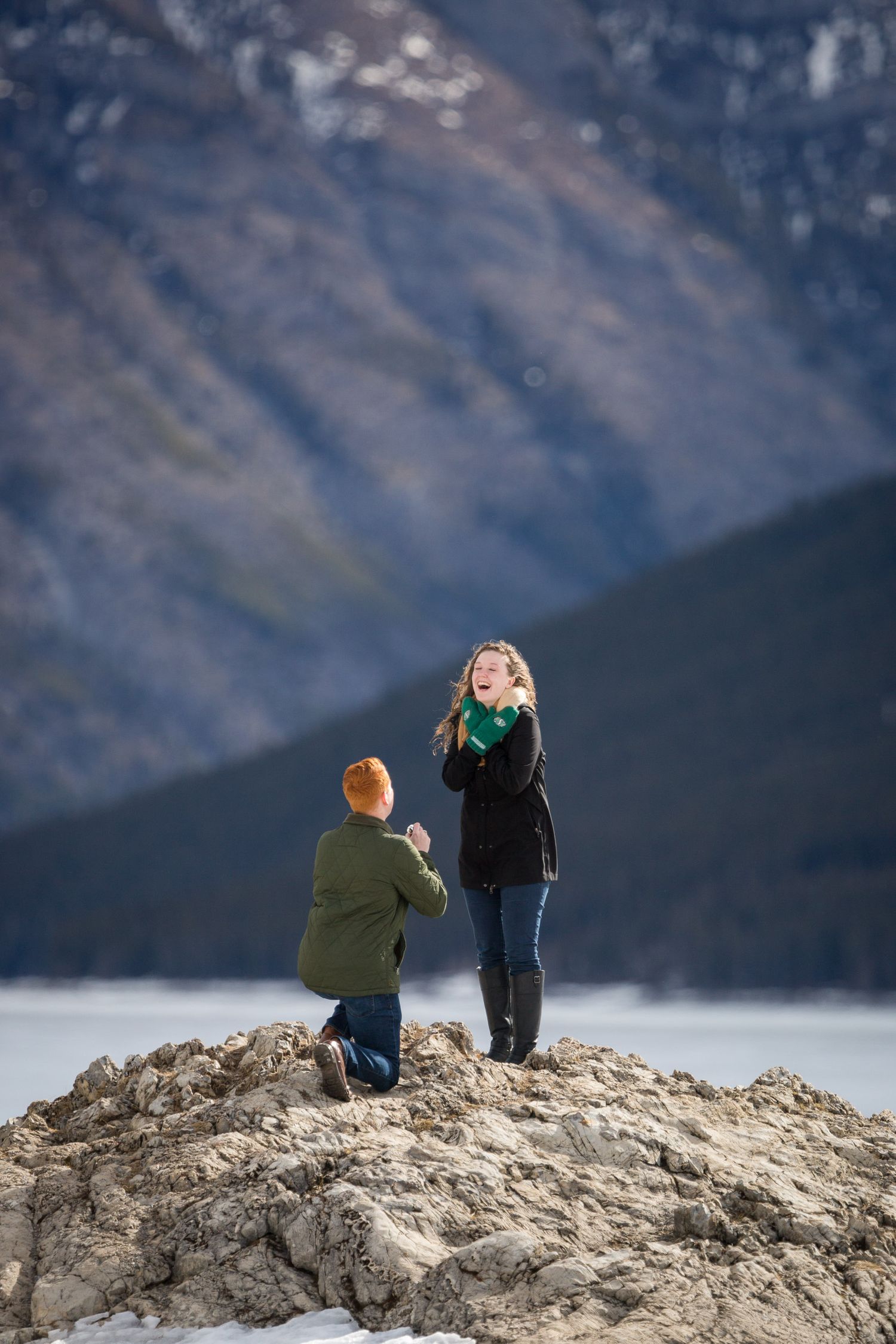 Man proposing on one knee to surprised woman on rocky outcrop with mountain views at Lake Minnewanka in Banff National Park.