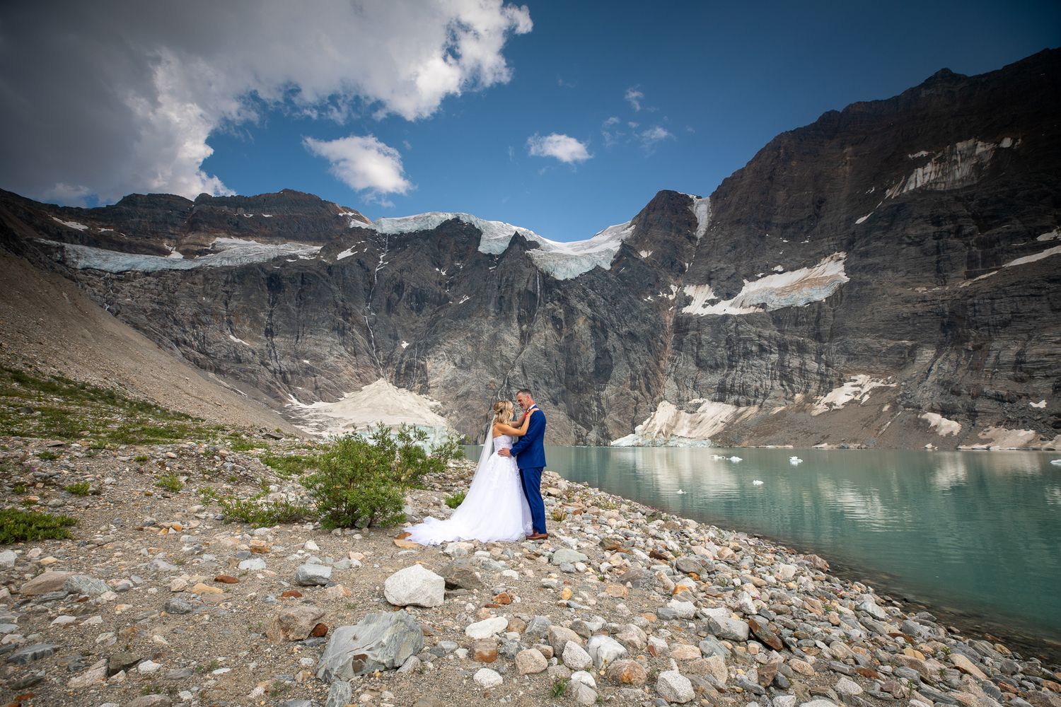 A bride and groom embrace before a stunning glacial lake and rocky mountain backdrop under a blue sky at Lake-of-Hanging-Glacier.