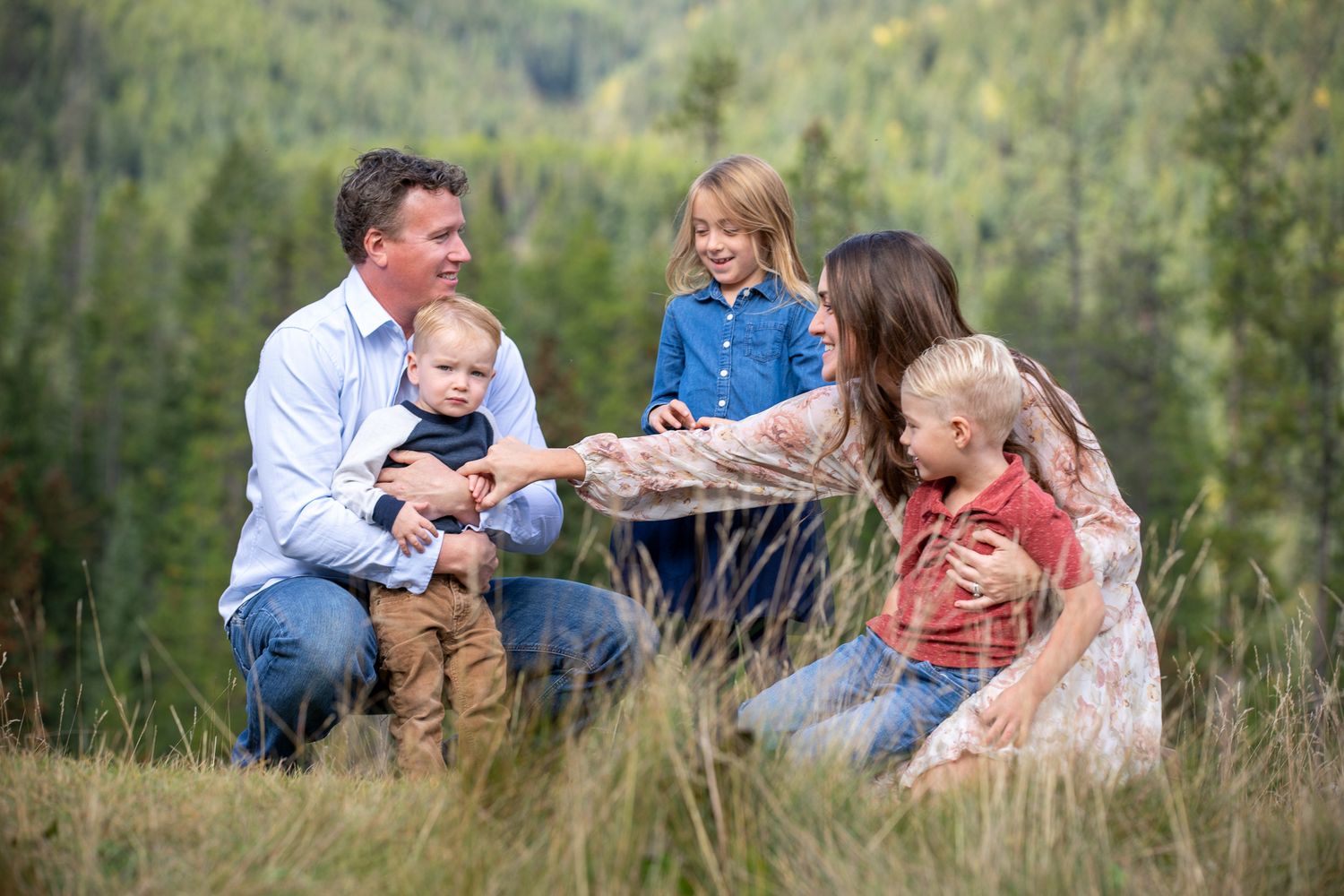 A group of people sit together in a grassy meadow surrounded by trees during an outdoor photo session at Grey Wolf in Panorama, BC.
