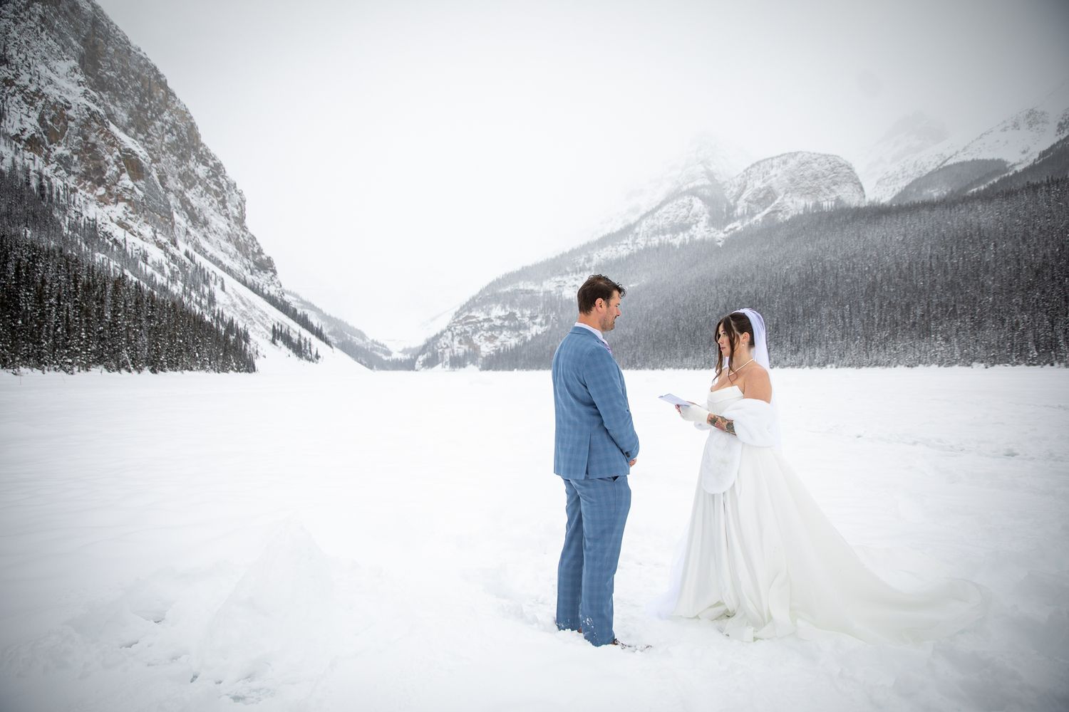 A couple shares an intimate moment during a wedding ceremony in a snowy winter landscape with mountain background at Lake Louise, Alberta.