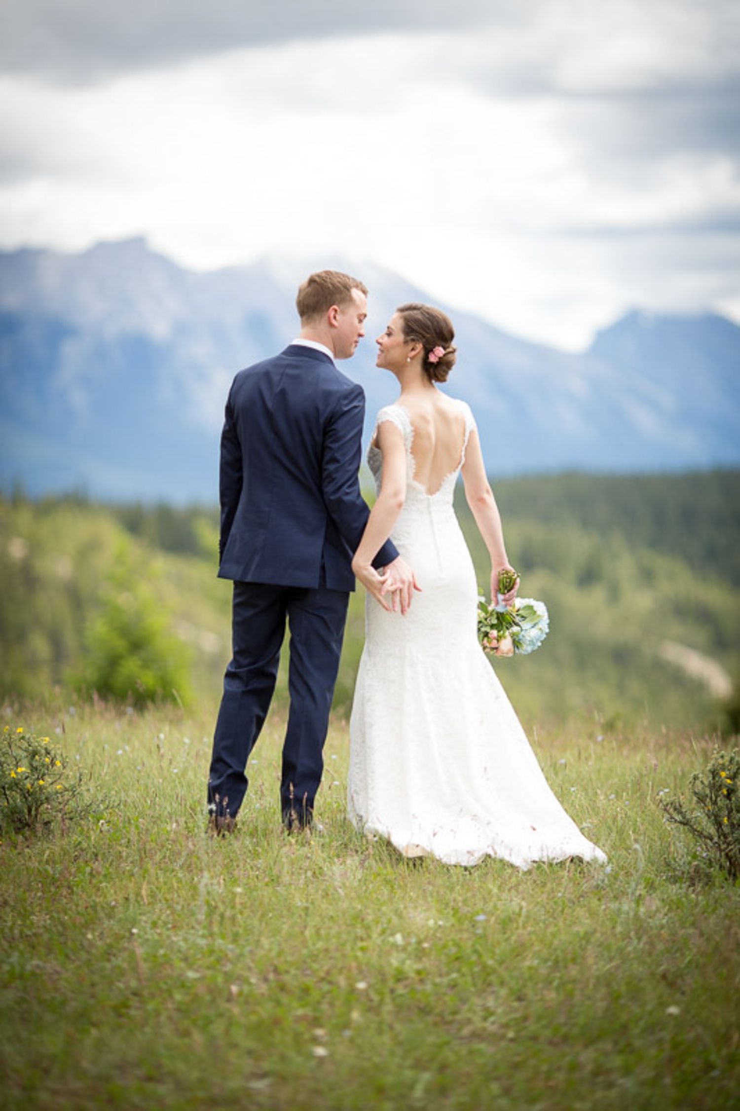 A couple in wedding attire walks together on a scenic mountain meadow with wildflowers and evergreen forests in the distance. Banff, Alberta
