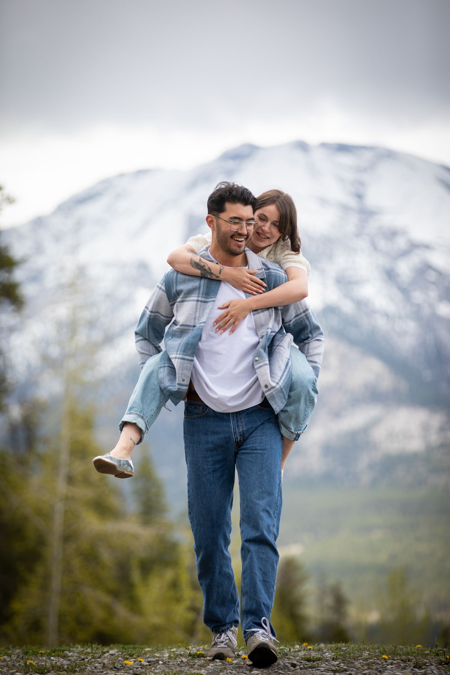 A couple shares playful moments against a backdrop of snow-capped mountains in casual denim outfits. Canmore Nordic Centre.