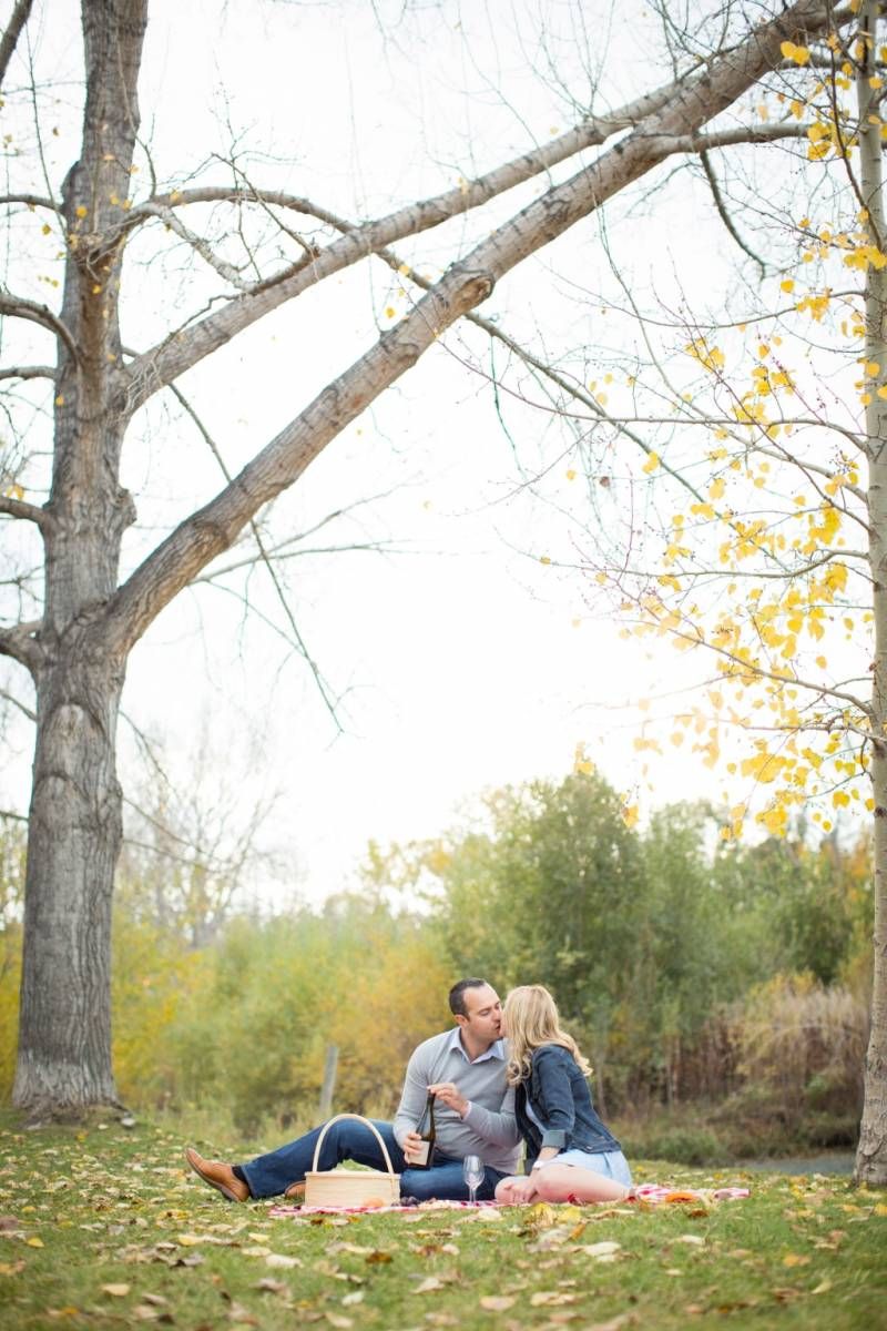 Couple kissing while sitting on picnic blanket under large trees with fall foliage at Fish Creek Park in Calgary.