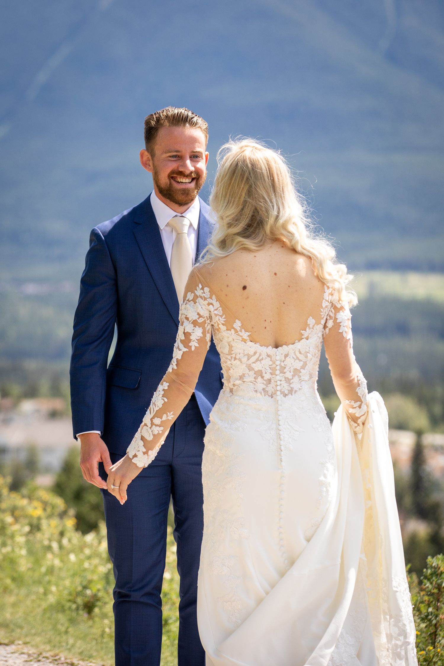 Smiling groom in navy suit holds bride's hand in lace wedding dress with mountain backdrop In Canmore Benchland.