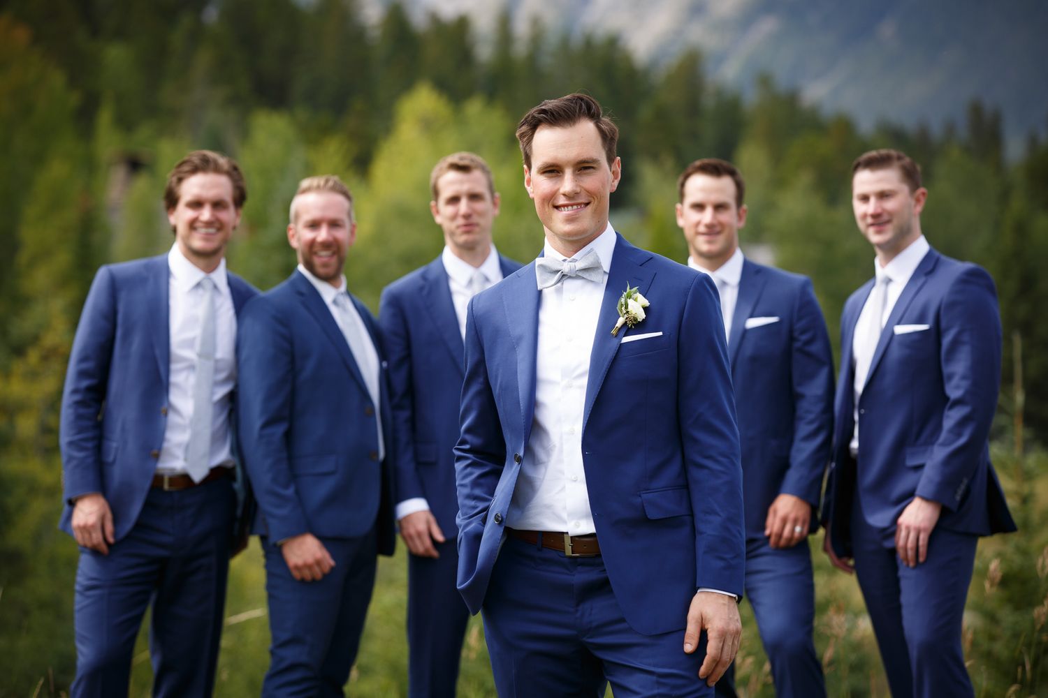 A group of groomsmen in navy blue suits pose together outdoors with mountain scenery and evergreen trees in background. Quarry lake park.