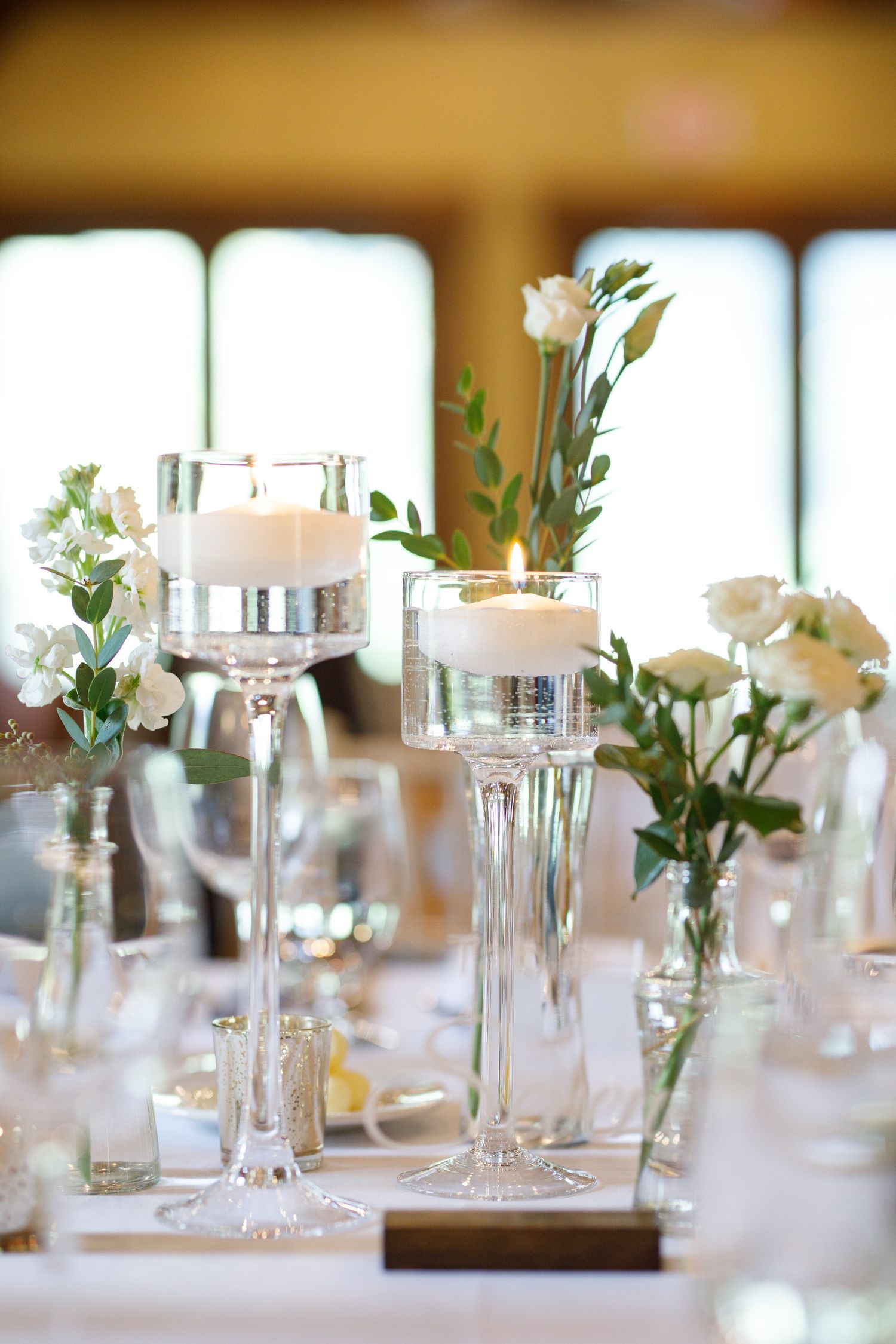 White floral centerpieces with glass vases and candles decorate elegant wedding reception tables in soft natural light.