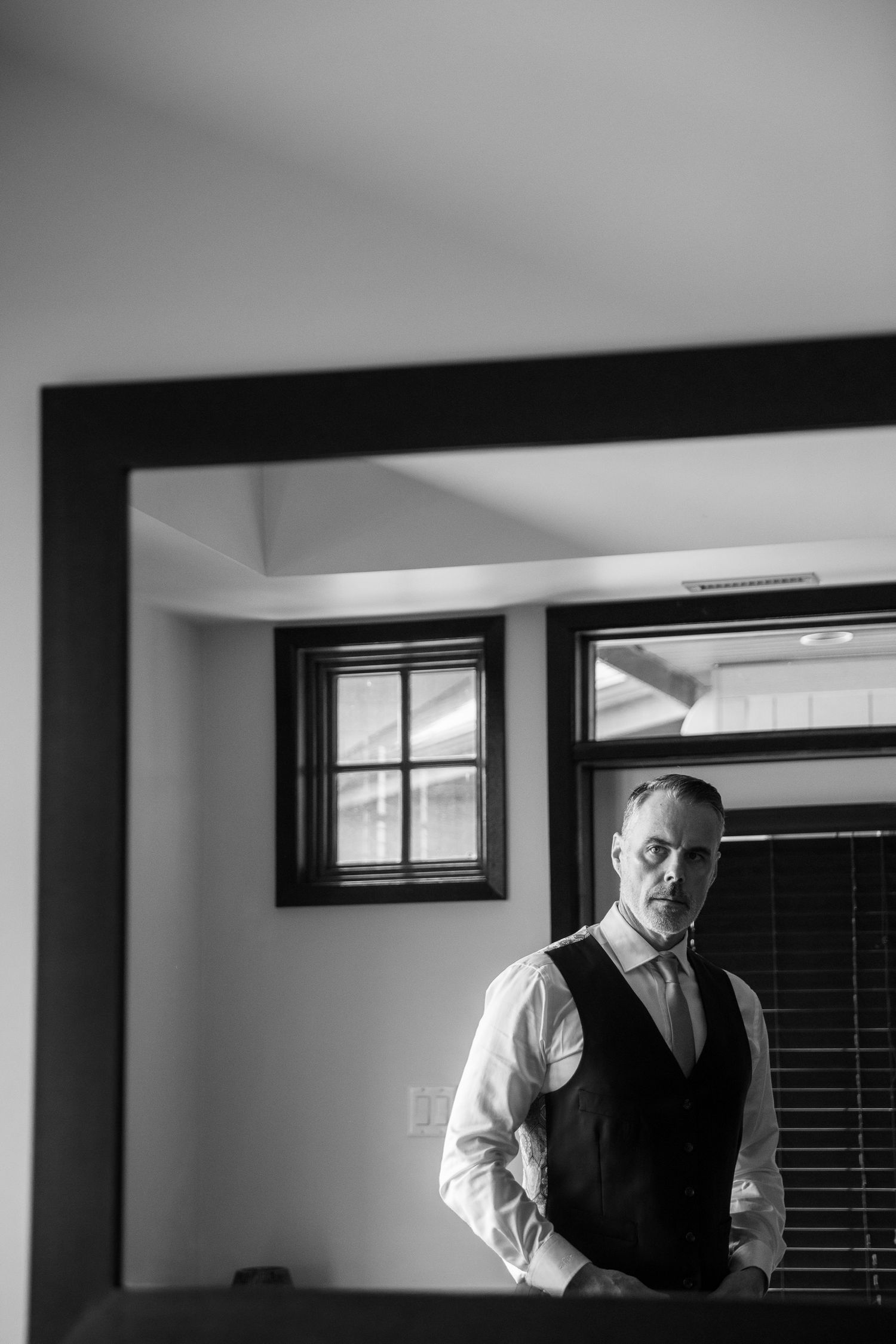 Black and white photo of a well-dressed man in vest and tie standing by window, reflected in a dark-framed mirror. Copperpoint resort.