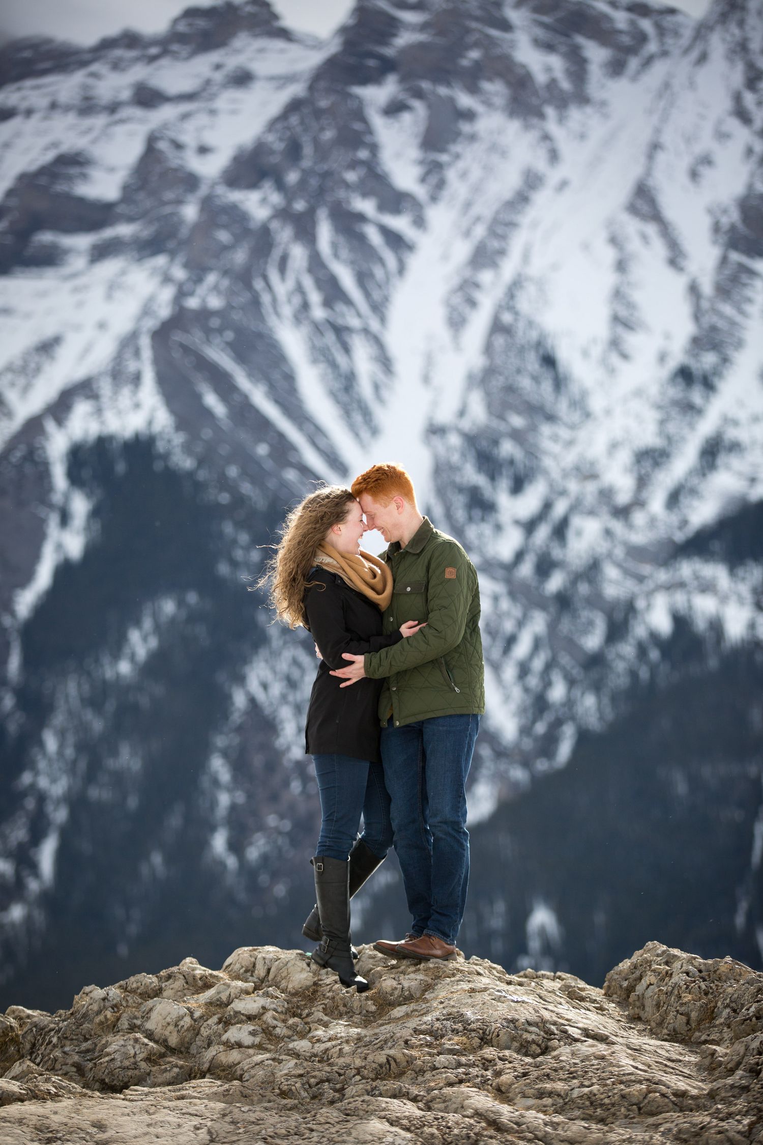 Couple kissing on rocky summit with snow-capped mountains backdrop in winter at Lake Minnewanka in Banff National Park.