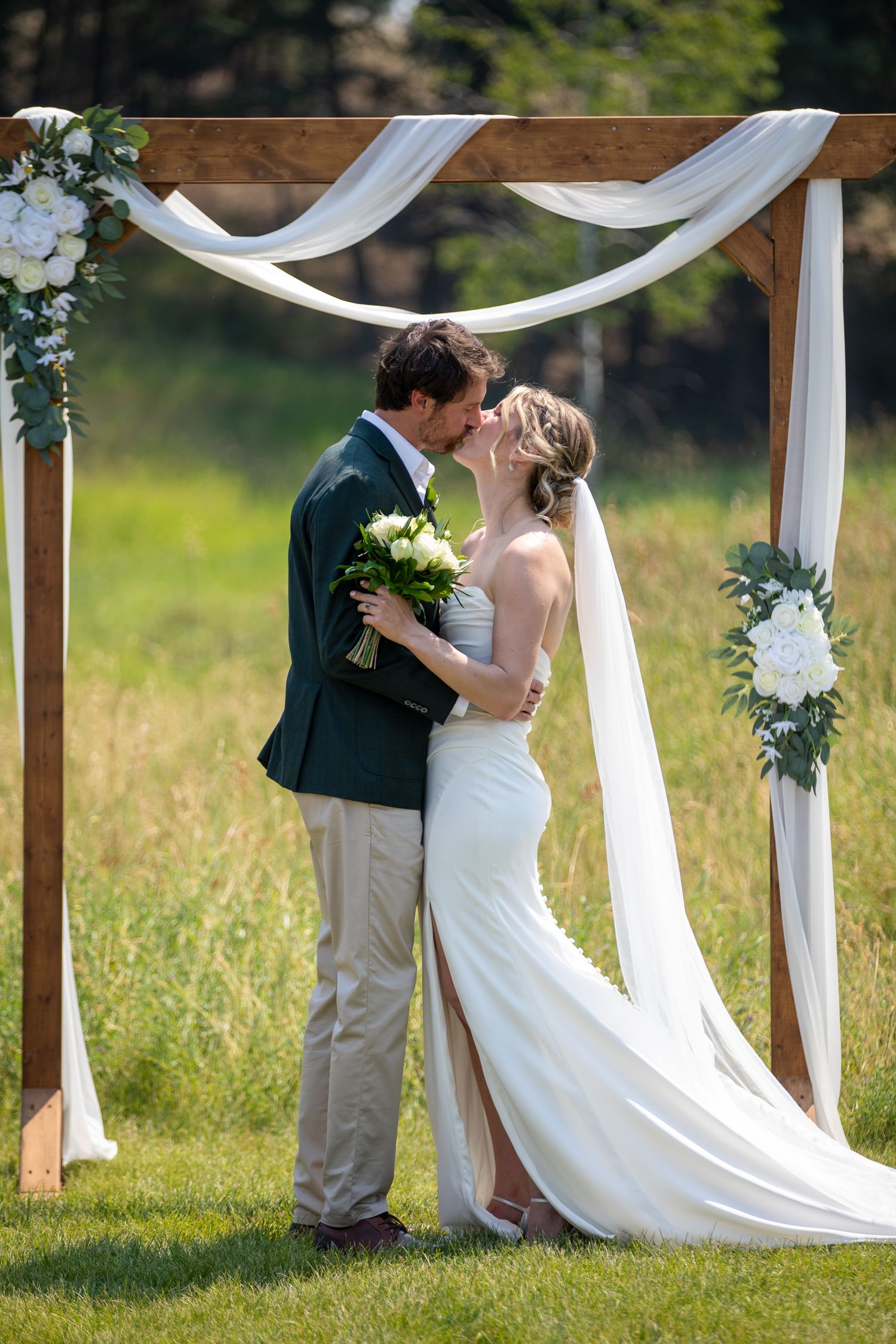 Bride and groom share first kiss under rustic wooden arch with white draping and floral accents at outdoor wedding at Swansea Ranch in BC
