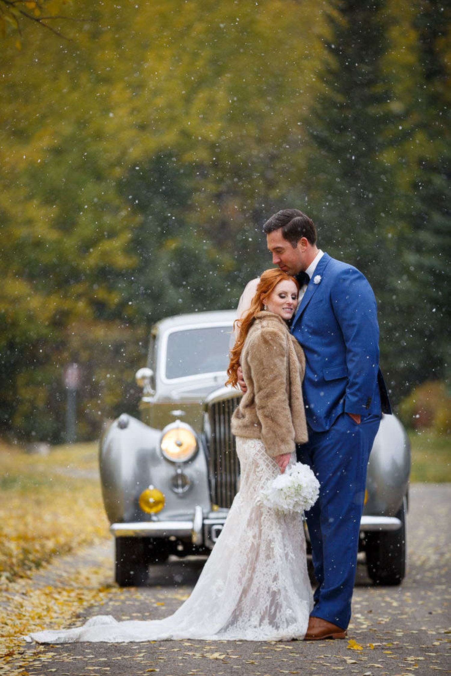 A romantic couple in formal attire embraces on an autumn road with a vintage car in the background during snowfall. Canmore Alberta.