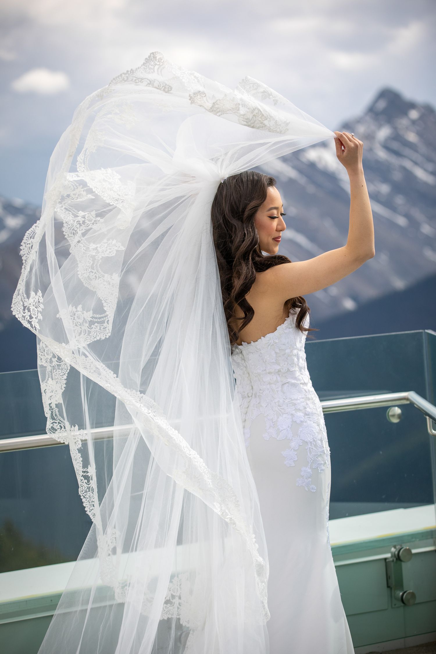 Bride in white lace gown with flowing veil billowing in wind, posing on mountain terrace with snow-capped peaks behind her.