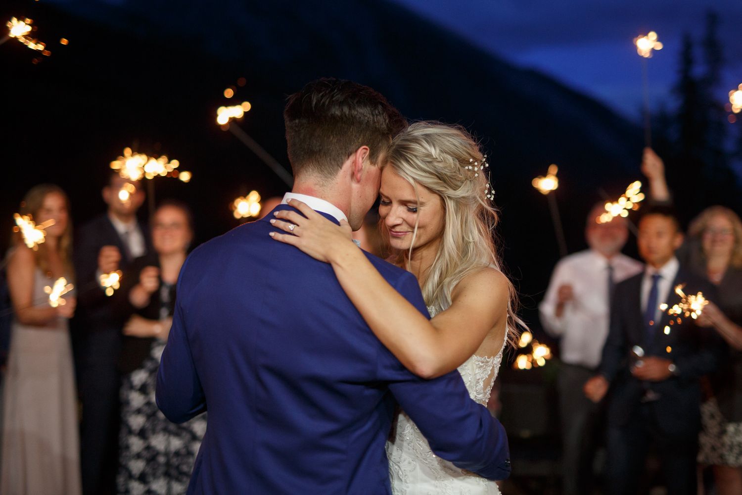A couple dances together at a wedding reception while holding sparklers as guests watch in the background. Silvertip resort in Canmore.