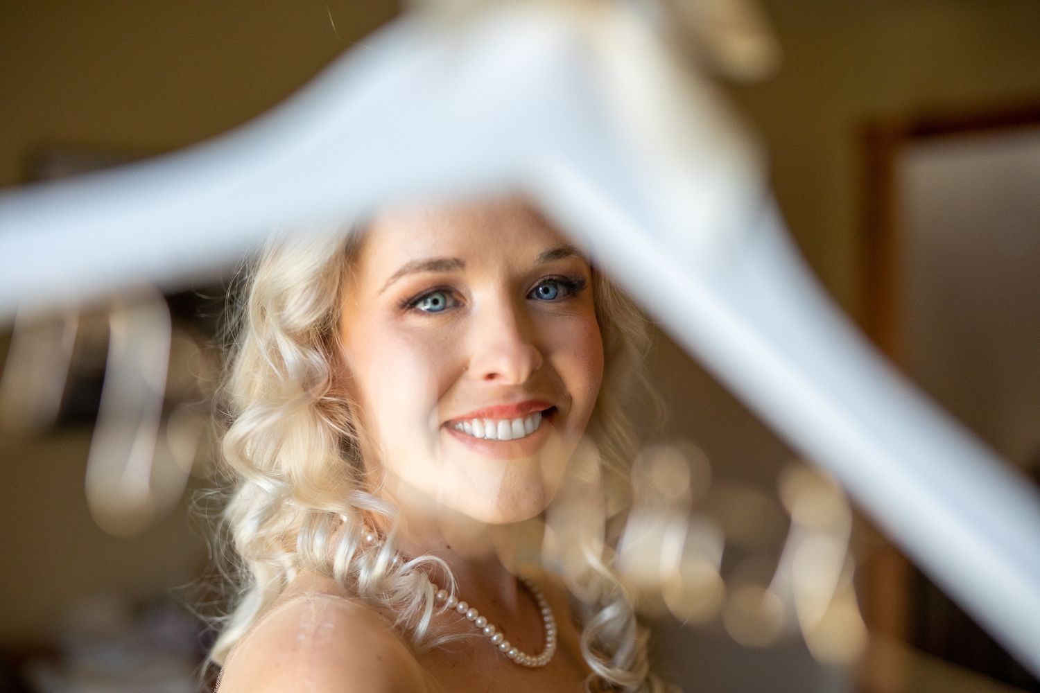 Smiling blonde bride wearing pearl necklace, framed by white wedding dress hanger, with soft bokeh background In Canmore