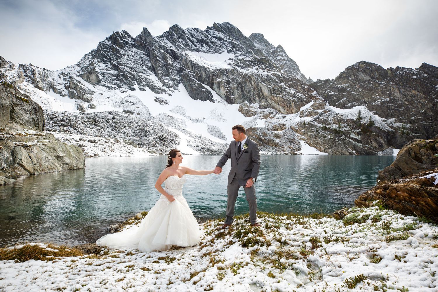 Bride and groom hold hands near a snowy alpine lake with dramatic mountain peaks in a stunning winter wedding photo in Helicopter wedding