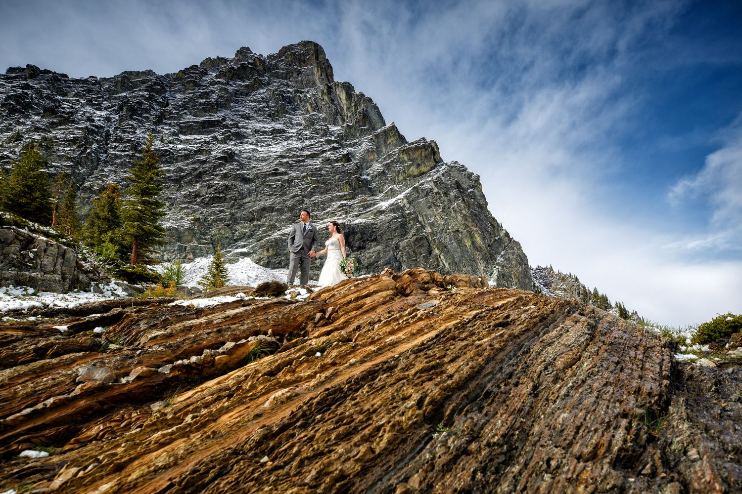 Bride and groom stand on layered rock formations with a dramatic mountain peak and blue sky backdrop. Cranbrook backcountry