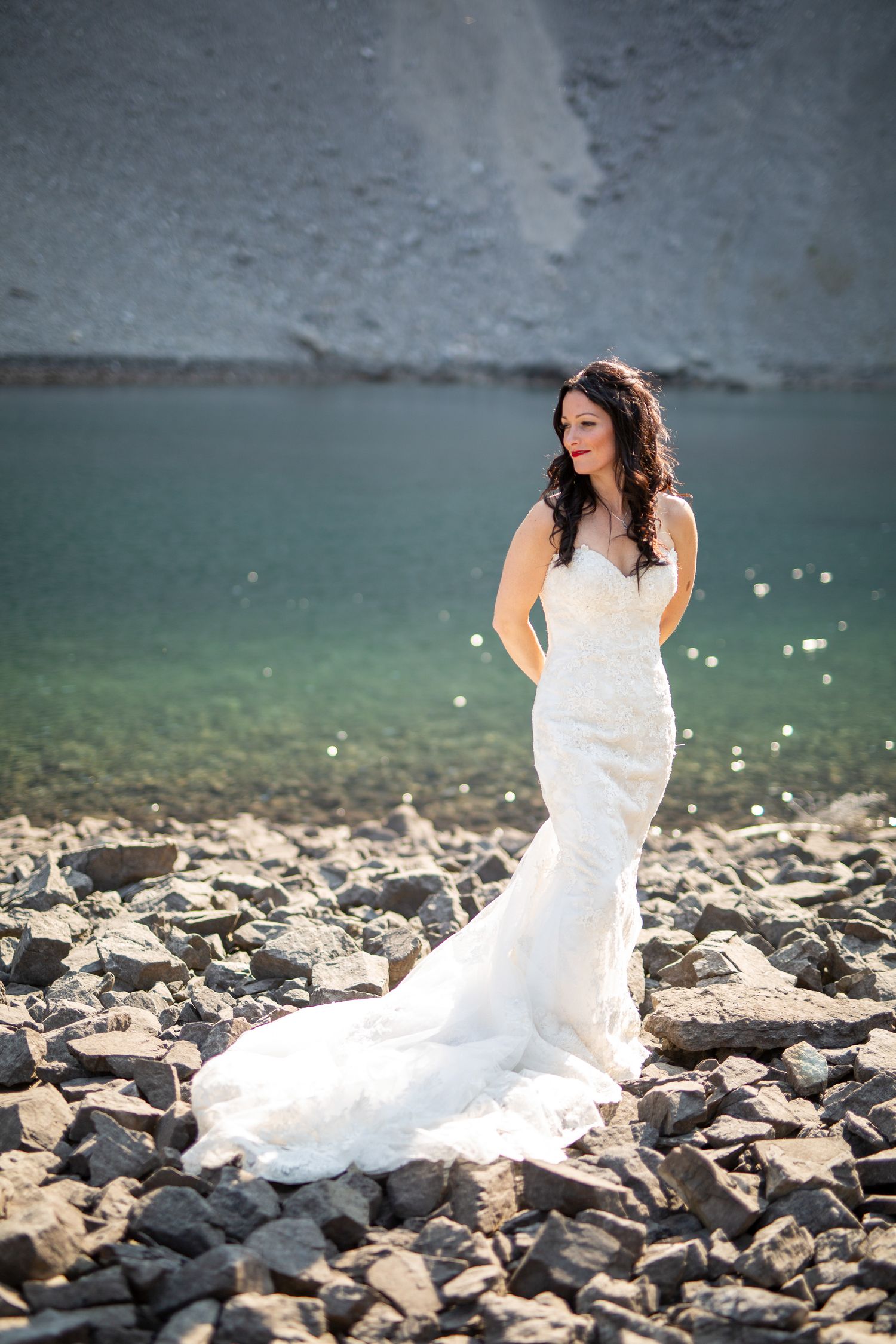 Bride in white mermaid gown poses on rocky shoreline with turquoise water and cliffs in background in Kananaskis, Alberta.