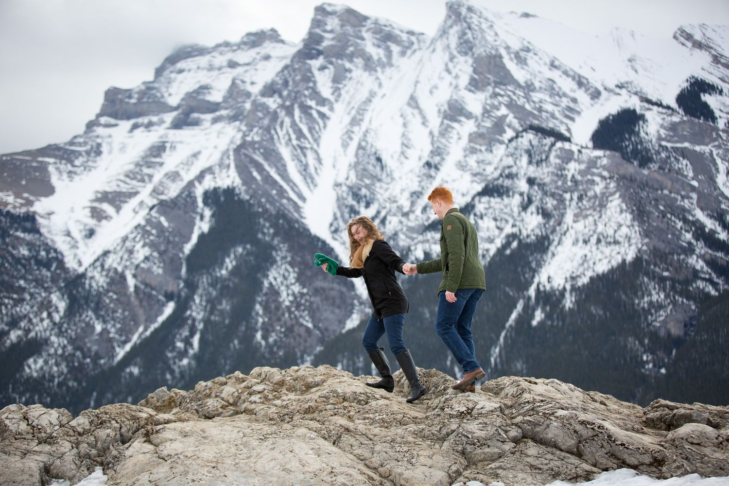Couple holding hands on rocky mountain summit with snow-capped peaks in background during winter at Lake Minnewanka in Banff National Park.