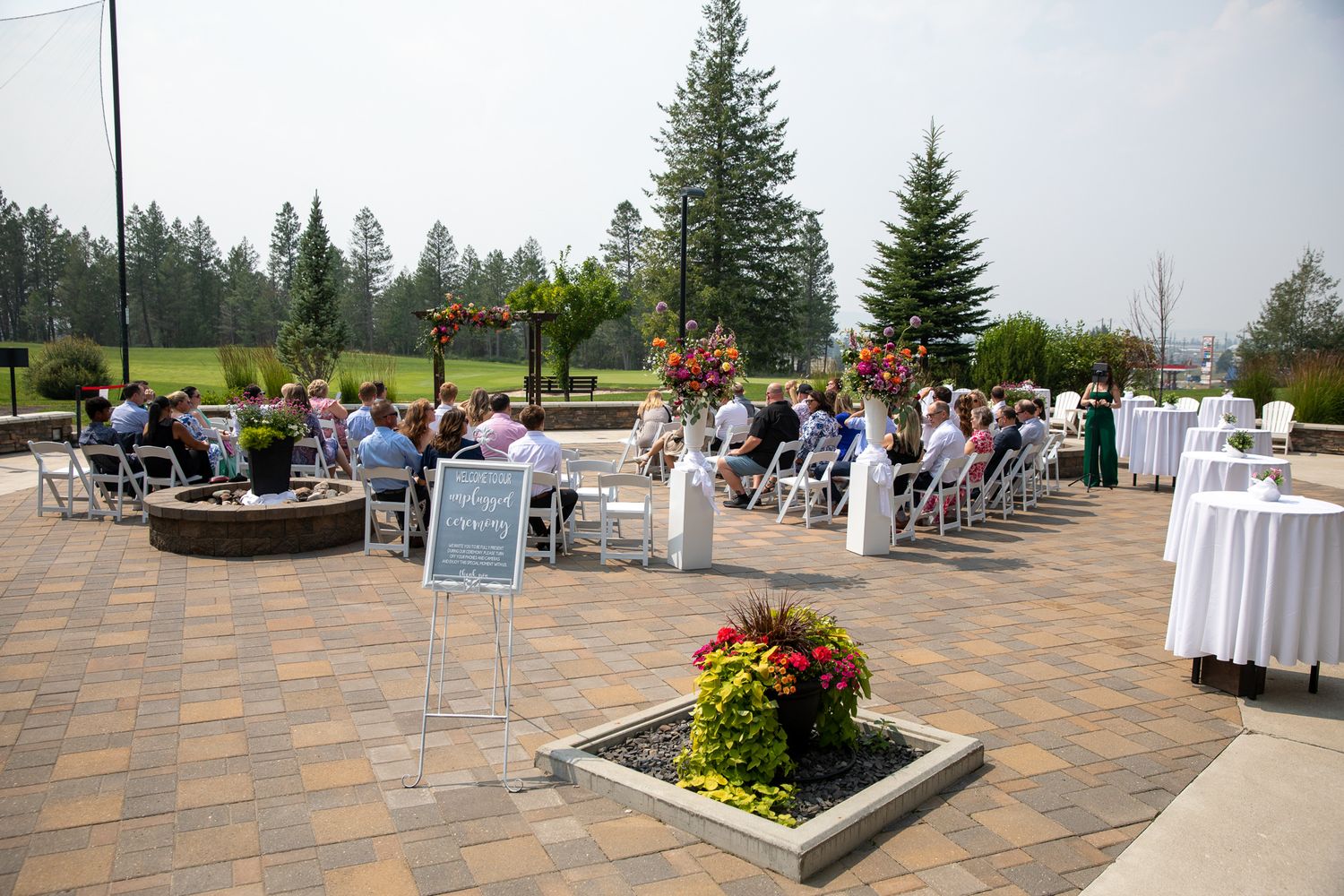 Outdoor wedding ceremony on a brick patio with white chairs, floral arrangements, and tall pine trees under a sunny sky. Copperpoint resort,