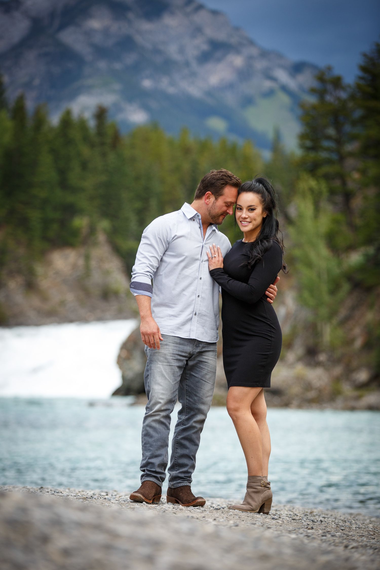 A couple embracing on a beach with a mountain landscape in the background at Bow Falls in Banff.