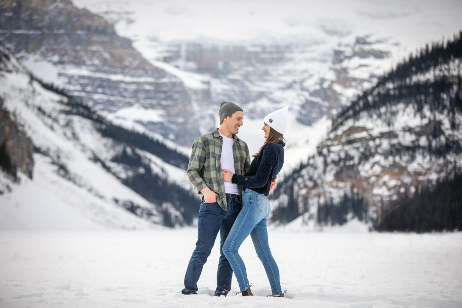 A couple in winter coats share a romantic moment in the snowy mountains of Lake Louise, Alberta.