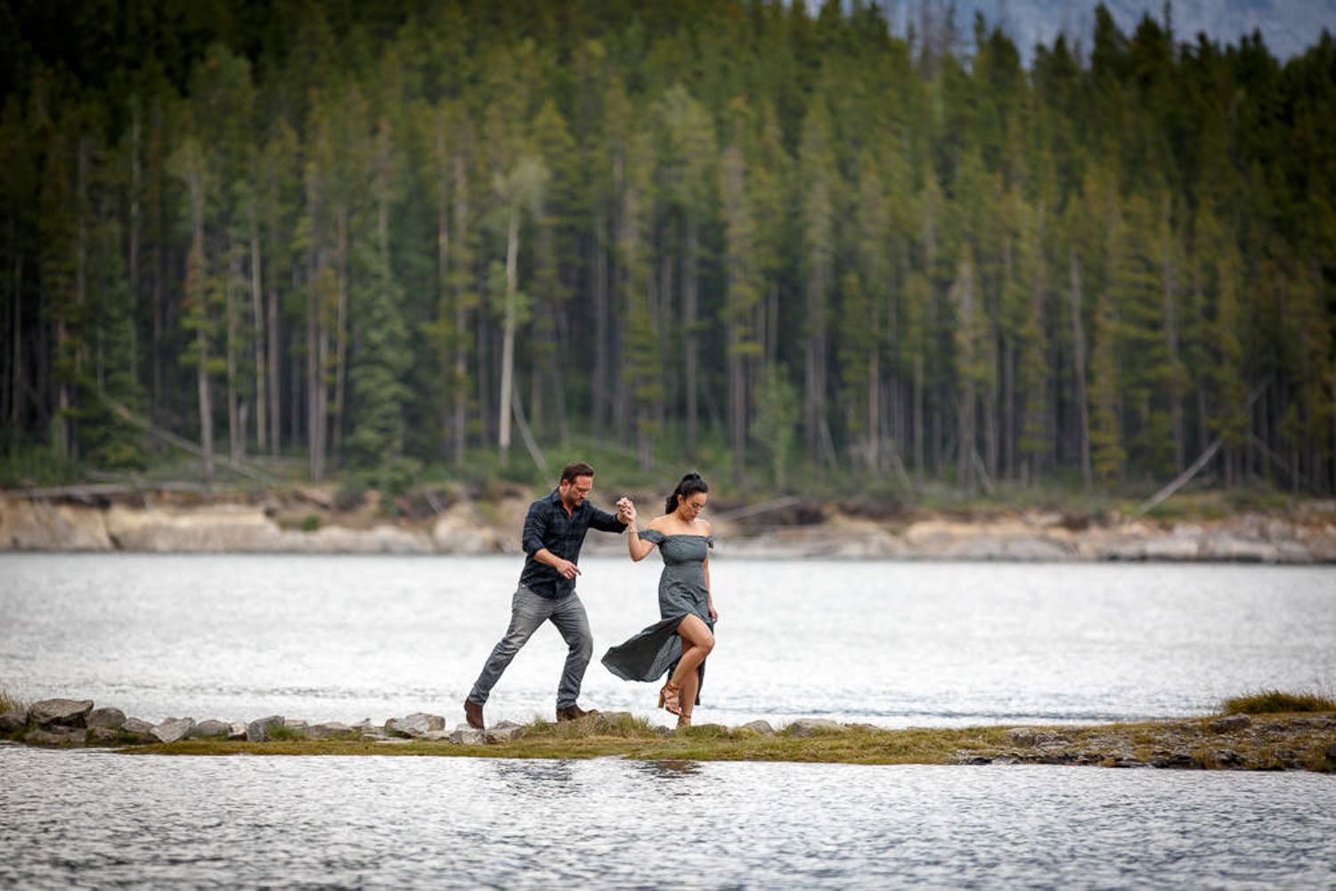 Two people dance together on a log by the water's edge with a forest backdrop at dusk at Lake Minnewanka in Banff.