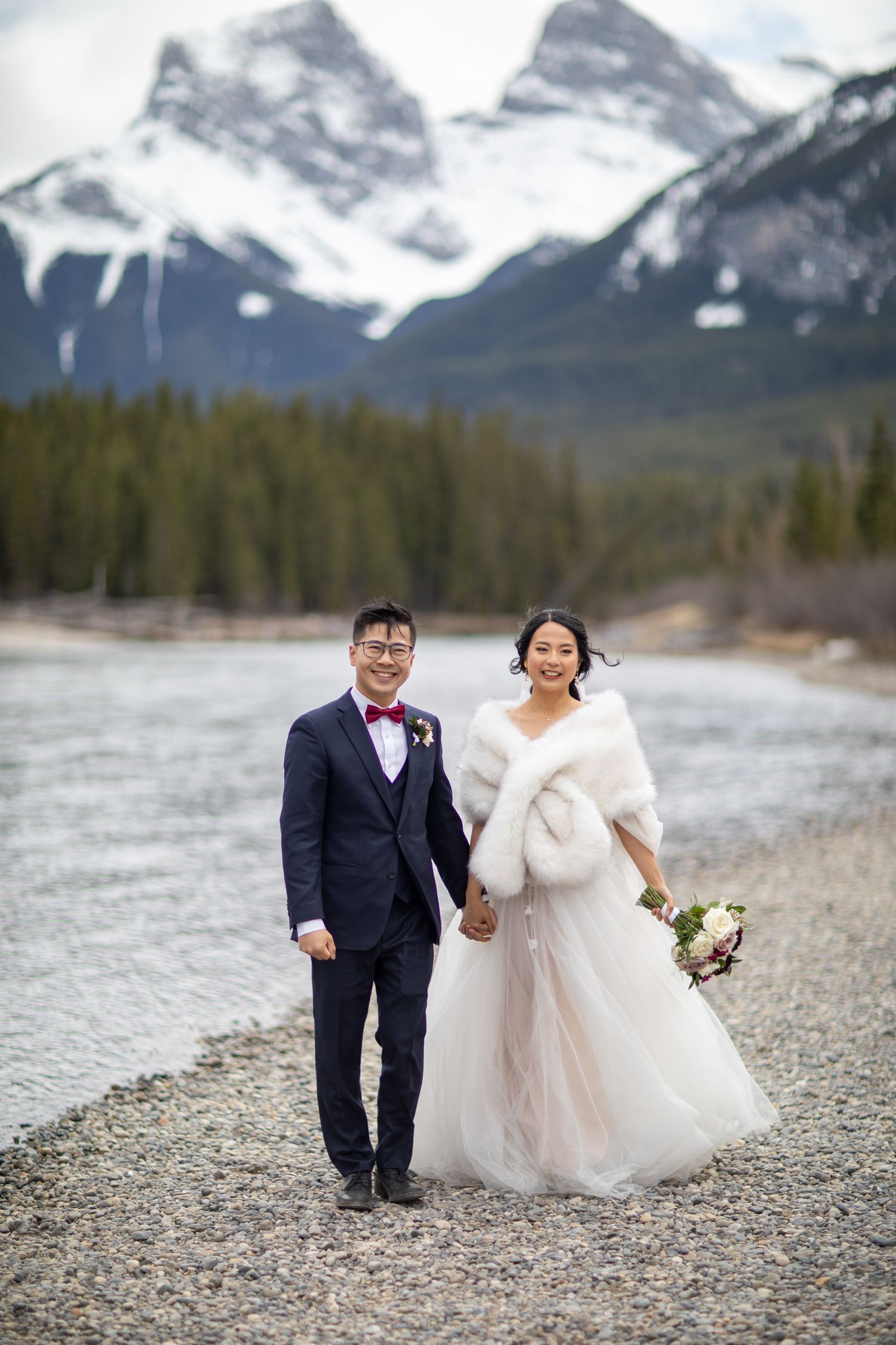 A couple in wedding attire walks along the Bow river with Three Sisters mountains in the background in Canmore, Alberta.