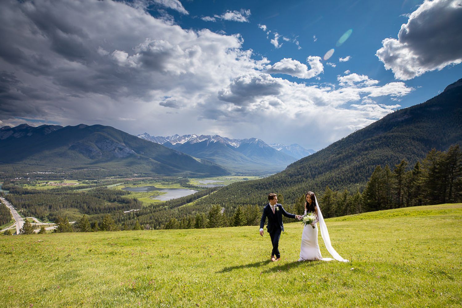 A scenic mountain wedding photoshoot in a lush green meadow with dramatic cloudy skies and snow-capped peaks. Mount Norquay lookout in Banff