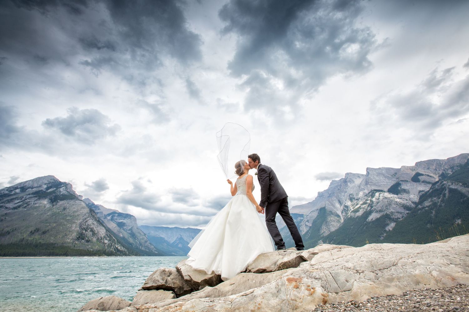 A wedding couple embraces on a rocky mountain overlook with dramatic cloudy skies and turquoise waters of Minnewanka lake in Banff, Alberta.