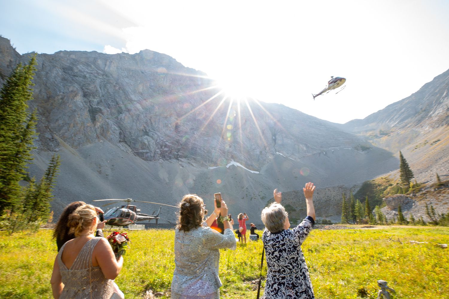 People waving at helicopters in a sunny mountain meadow with wildflowers during an outdoor celebration in Kananaskis, Alberta.