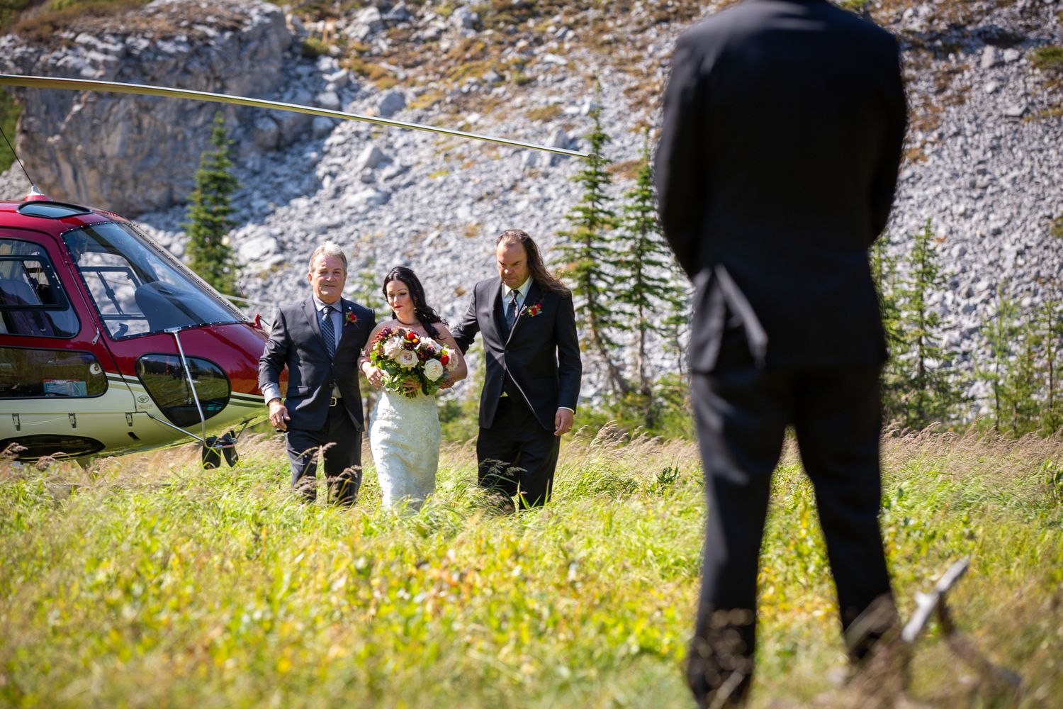 Bride walks toward groom with officiant near a red helicopter on a scenic mountain meadow during an outdoor elopement in Kananaskis, Alberta