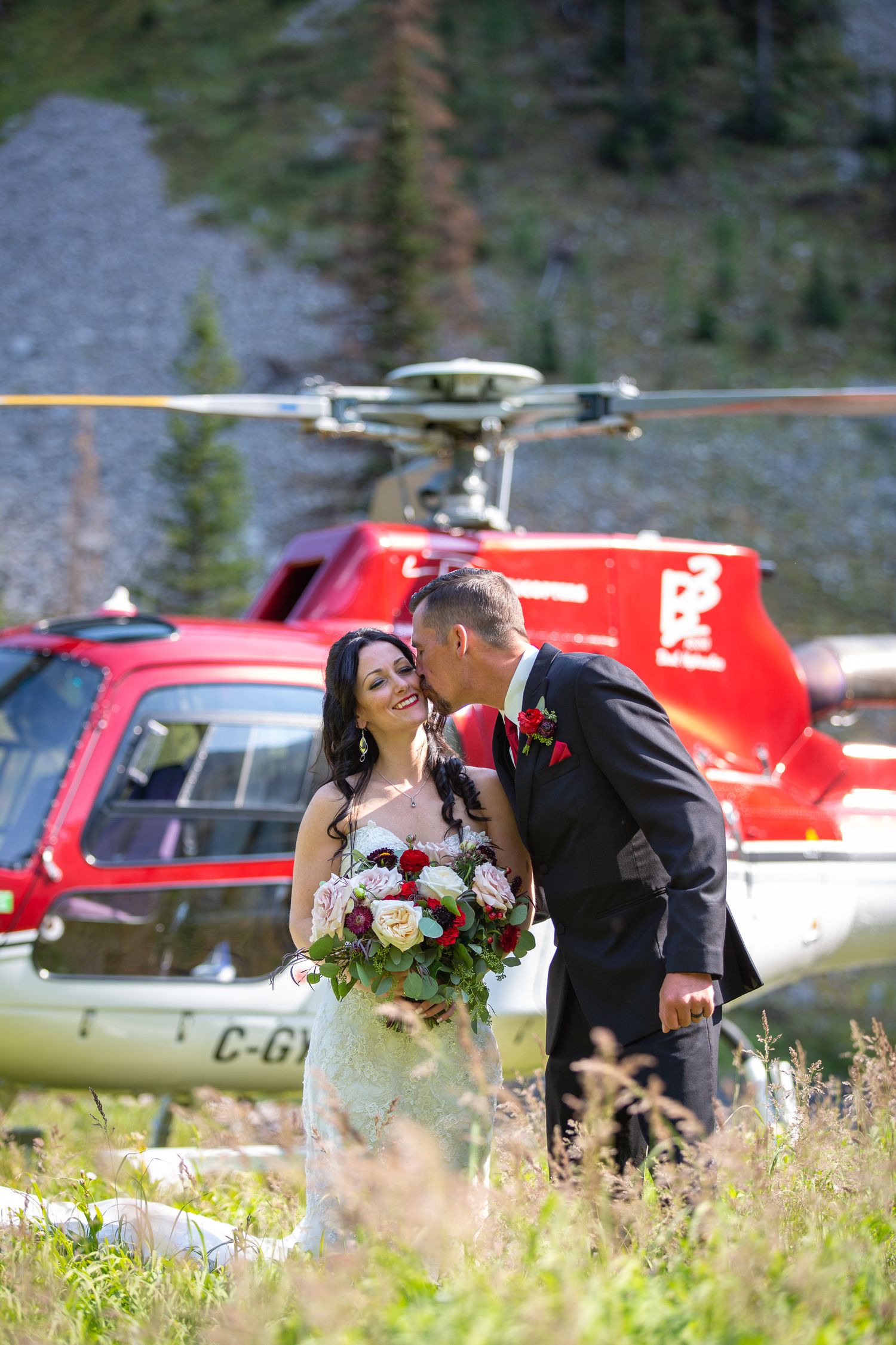 Bride and groom share a romantic kiss in front of a red helicopter on a mountain meadow wedding day in Kananaskis, Alberta.