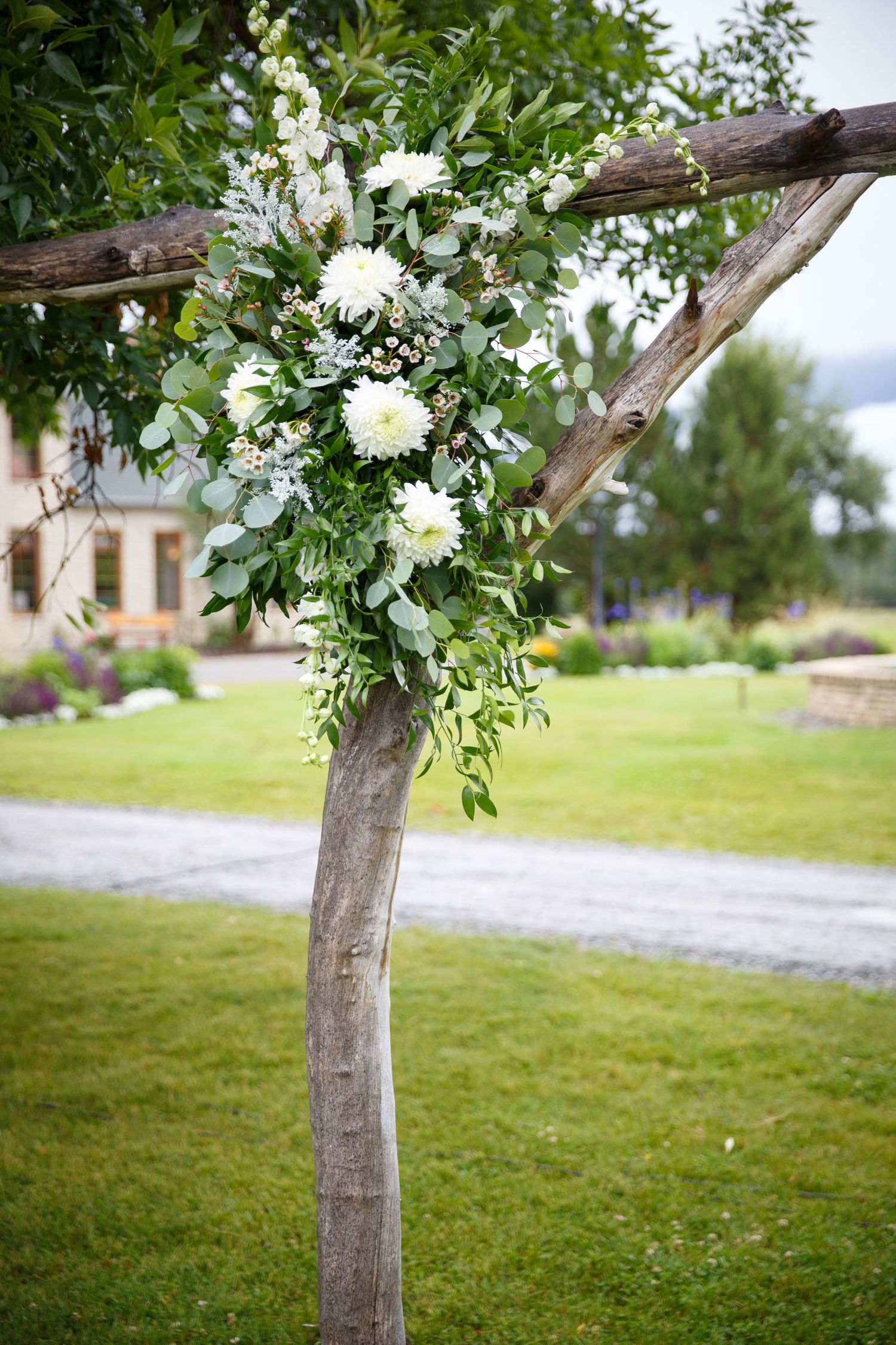 Rustic wooden wedding arch adorned with white dahlias and lush greenery, set against a manicured lawn outdoor venue.