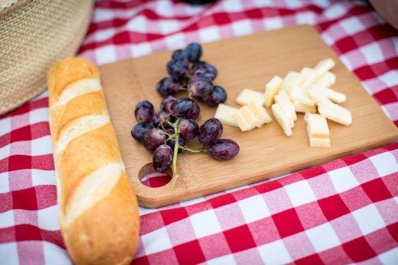 A wooden cutting board with grapes, cheese, and bread on a red checkered picnic blanket at Fish Creek Park in Calgary.