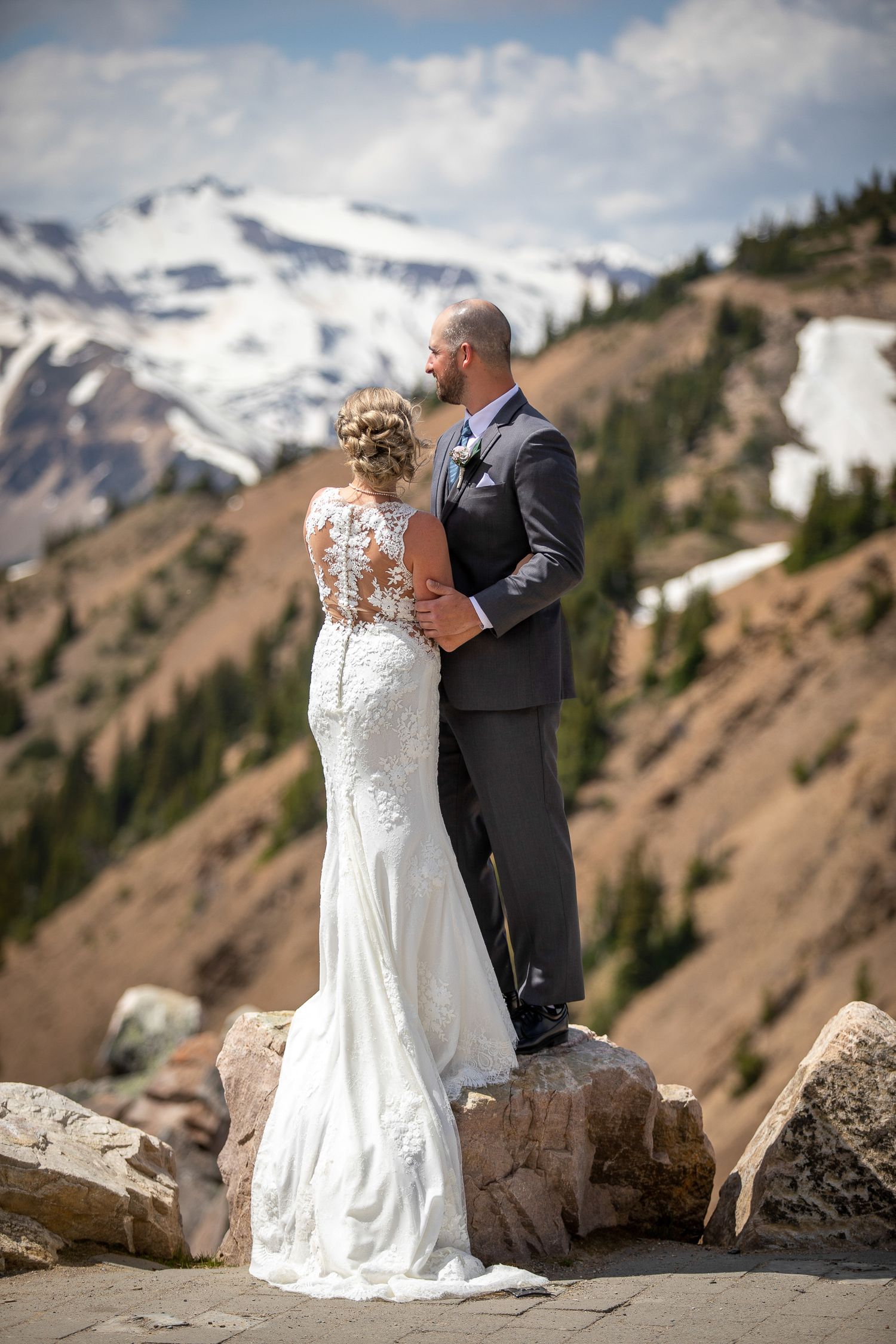 A romantic wedding couple embraces on a mountain peak with snow-capped peaks and rugged terrain in the background. Golden, BC
