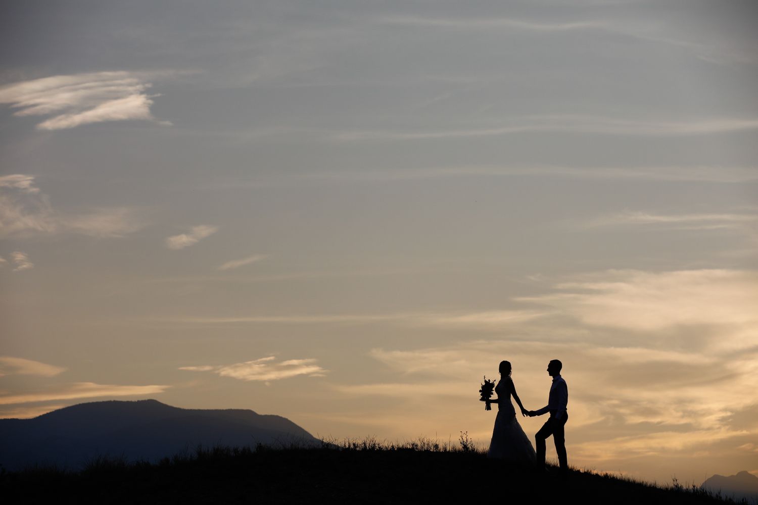 Silhouettes of bride and groom walking along a ridge at sunset with dramatic orange and pink sky at Swansea Ranch in Windermere, BC.