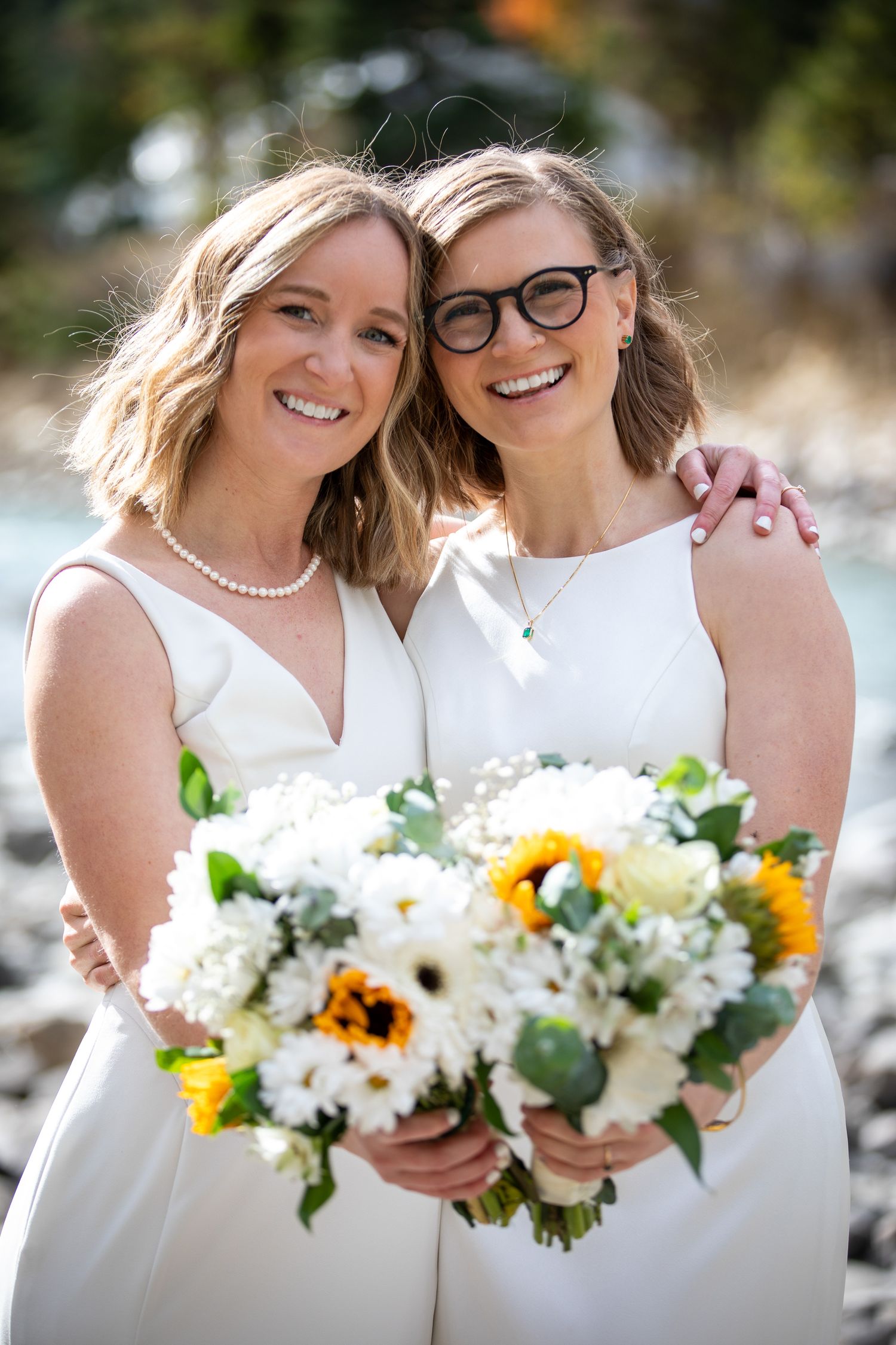 A LGBTQ in white wedding attire share joyful moments while holding beautiful bouquets of sunflowers and white blooms outdoors.