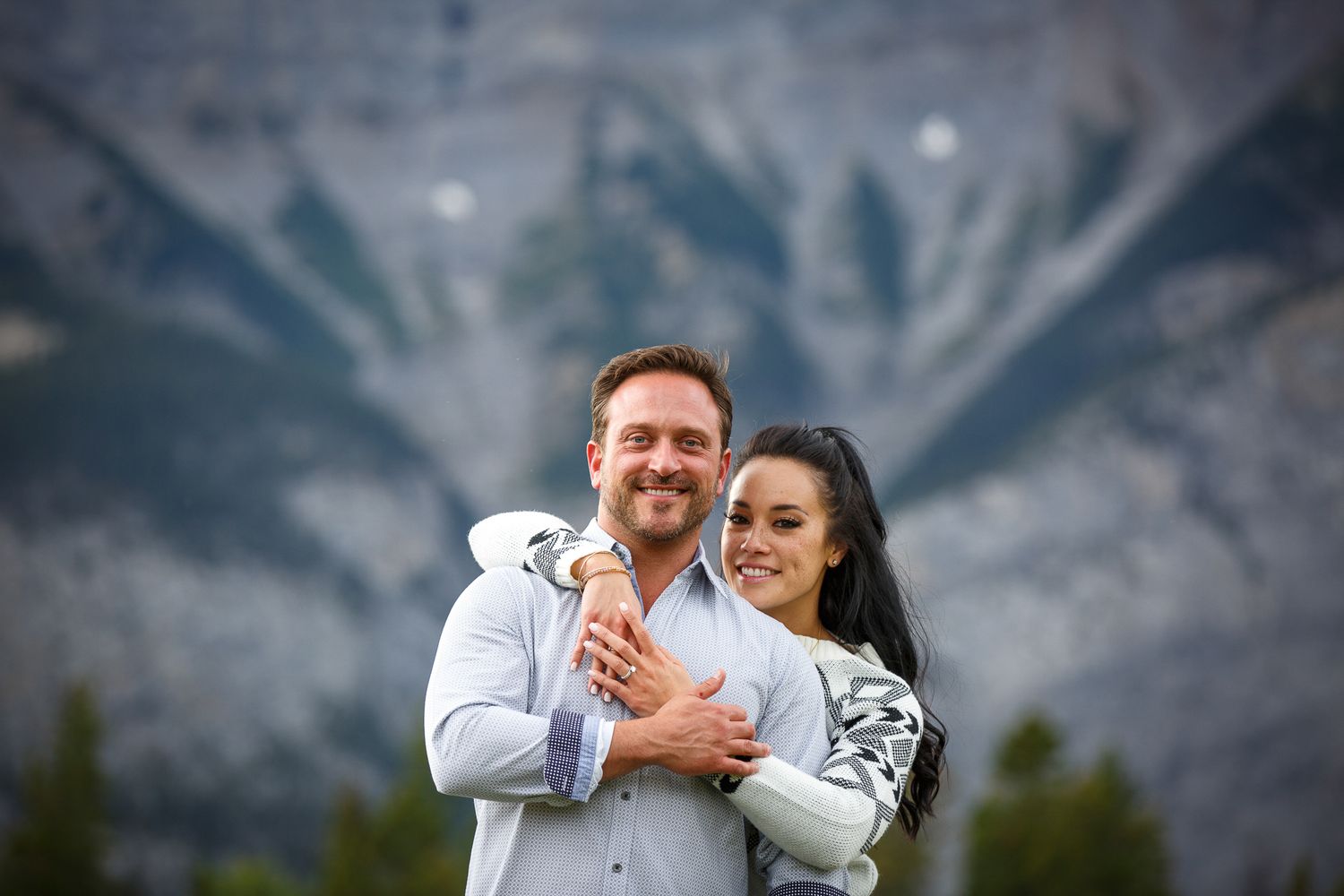 A smiling couple embracing outdoors against a snowy mountain backdrop at Lake Minnewanka in Banff.