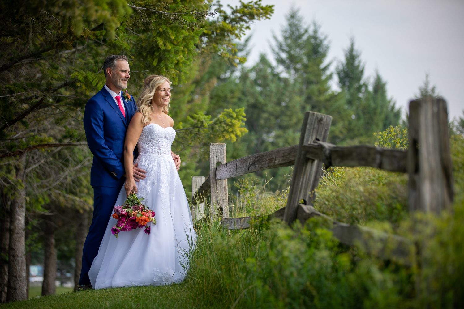 Bride and groom pose by rustic wooden fence surrounded by lush green trees on their wedding day at Eagle Ranch, Invermere, BC.